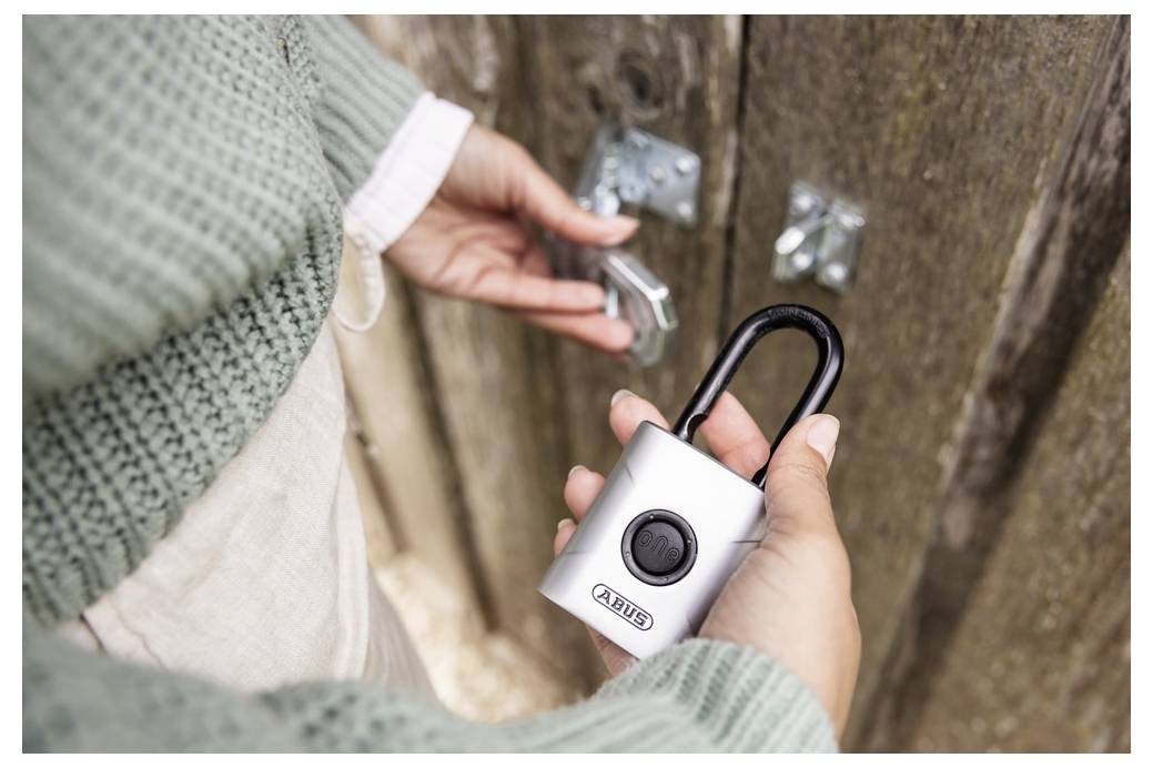 Une personne portant un pull vert déverrouille un cadenas sur une porte en bois à l'aide d'une clé. Le cadenas est argenté avec un logo de marque visible.
