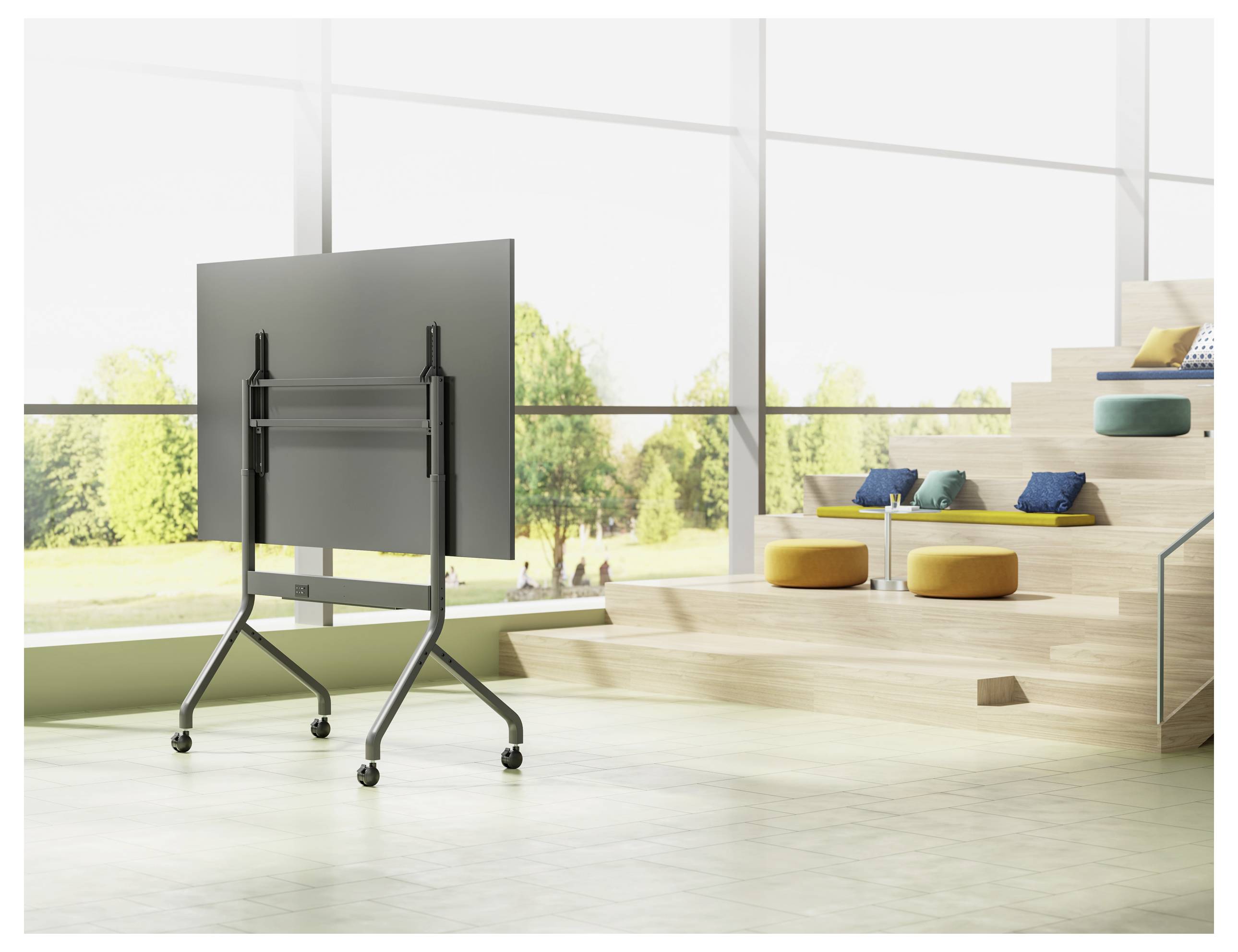 Une pièce spacieuse et lumineuse avec de grandes fenêtres, un écran de télévision mobile sur un support, et des sièges en gradins en bois avec des coussins colorés.