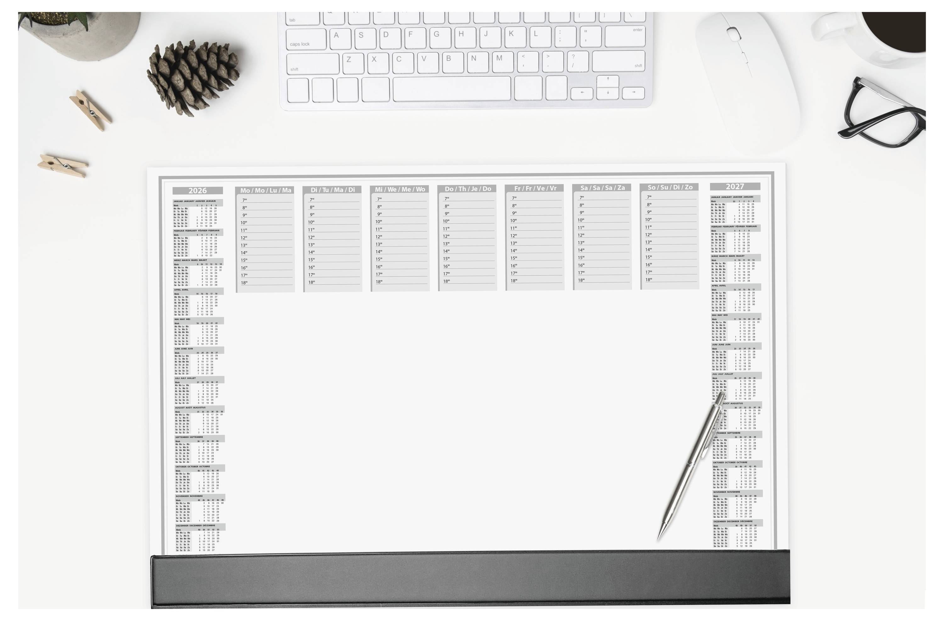 Un grand calendrier de bureau est posé sur une surface blanche, avec un stylo par-dessus. Un clavier, des lunettes, une pomme de pin et une tasse sont disposés à proximité.