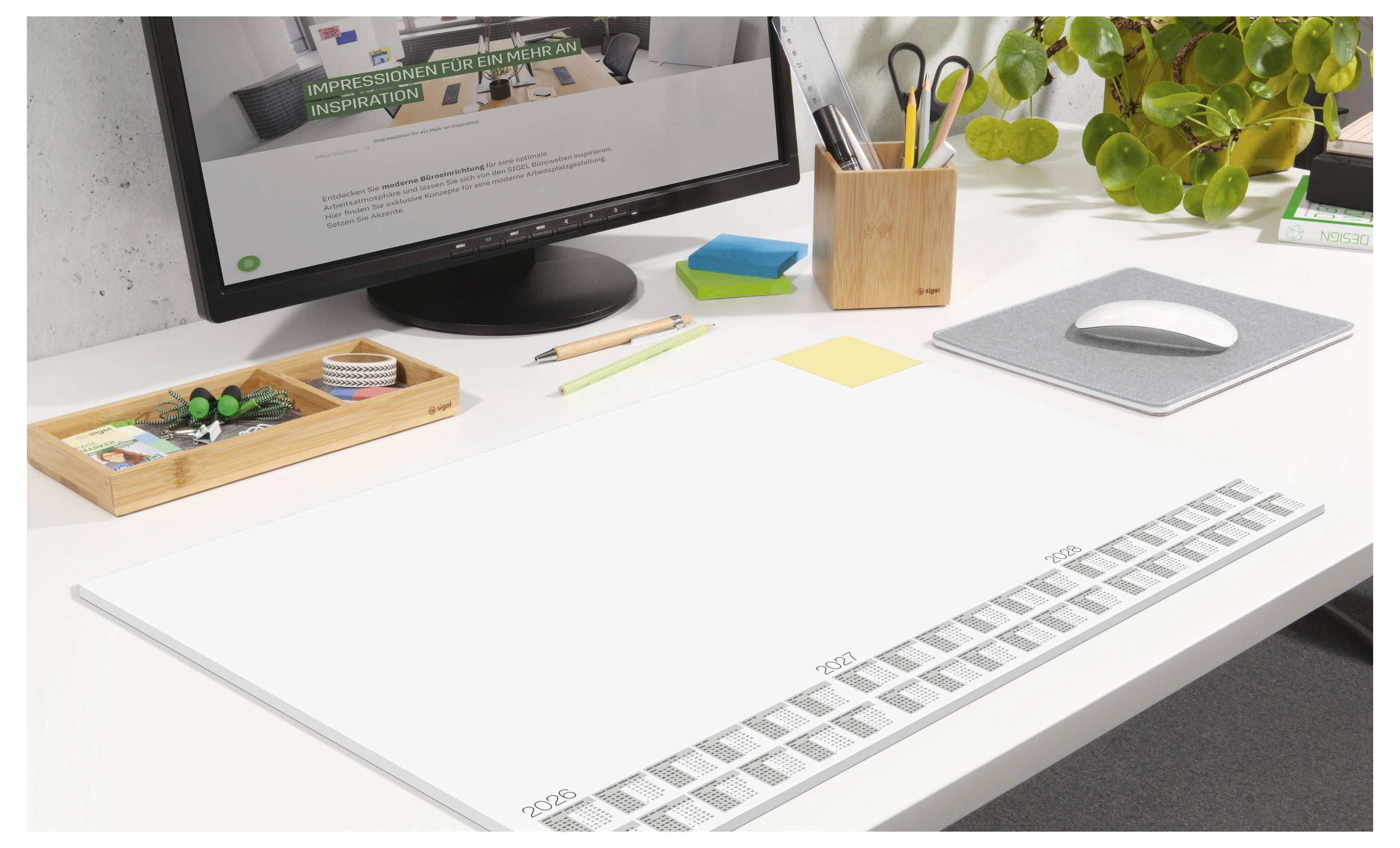 Un bureau de travail bien rangé avec un écran d'ordinateur, une souris, une plante, des stylos et un grand calendrier posé montrant les dates en bas.
