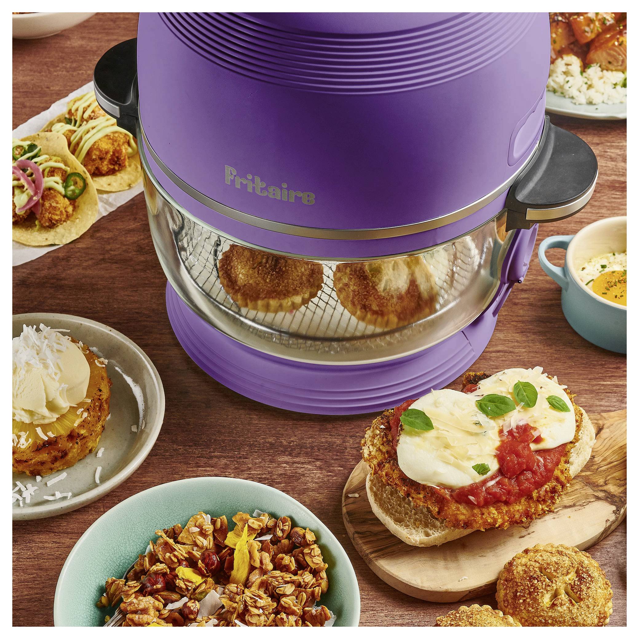 Un air fryer violet Fritaire avec deux muffins cuits à l'intérieur, entouré de différents plats dressés comprenant des tacos, une soupe et des pâtisseries sur une table en bois.