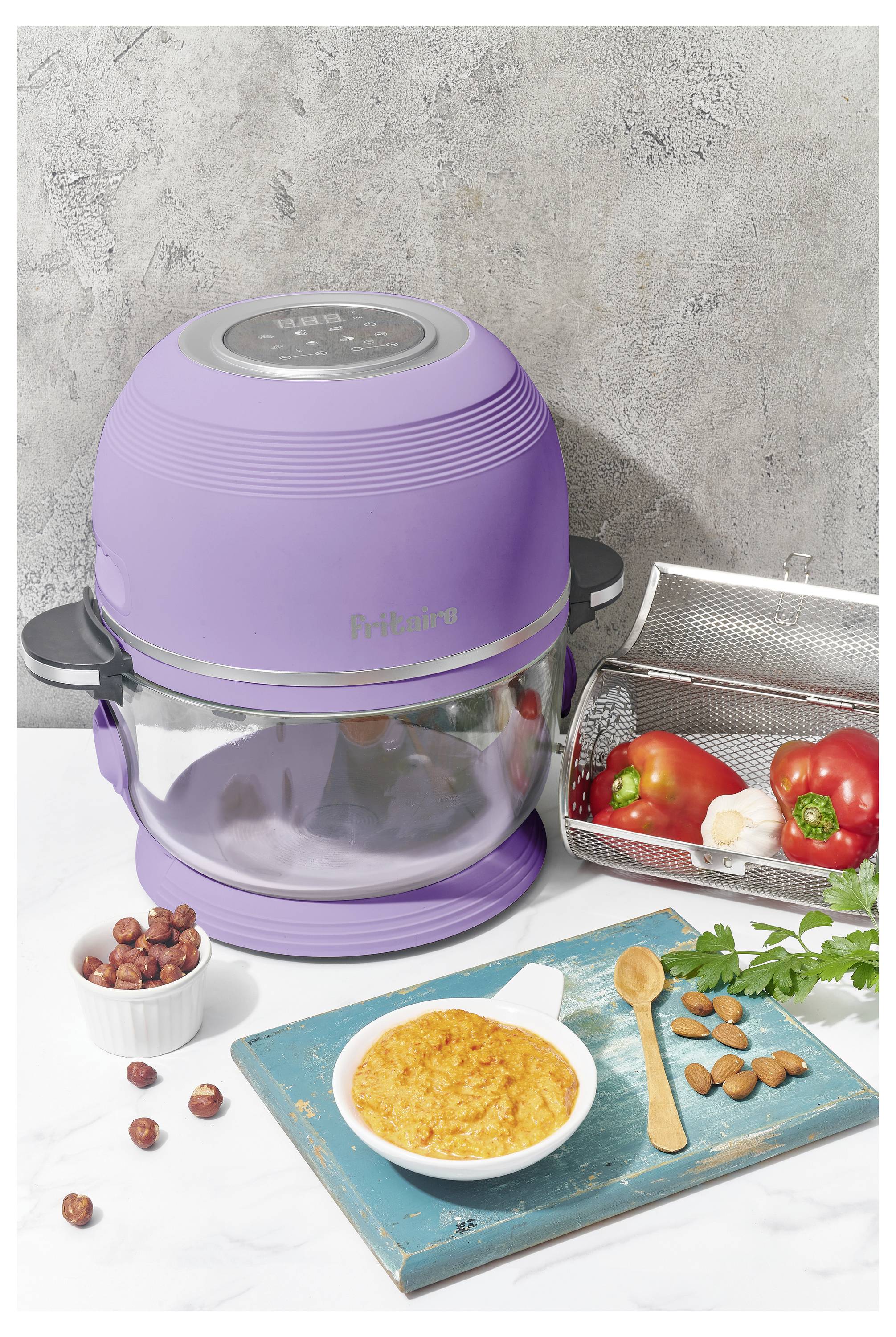 Un air fryer violet sur un plan de travail de cuisine avec un bol en verre. À proximité se trouvent un panier métallique rempli de légumes, un petit plat de noix et un bol de soupe.