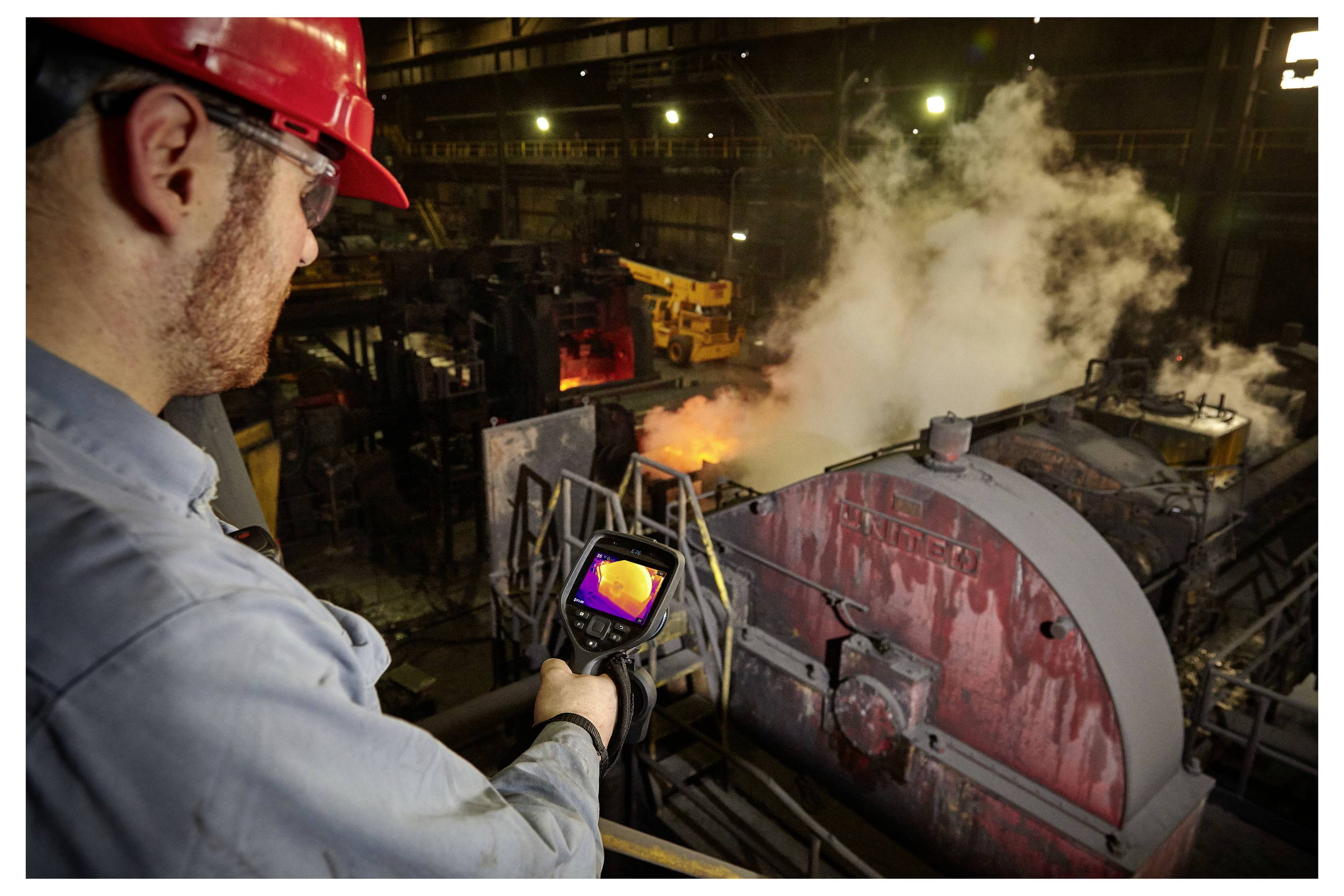 Un travailleur portant un casque de sécurité rouge utilise une caméra thermique pour surveiller des machines avec de la vapeur et des flammes dans un environnement industriel.
