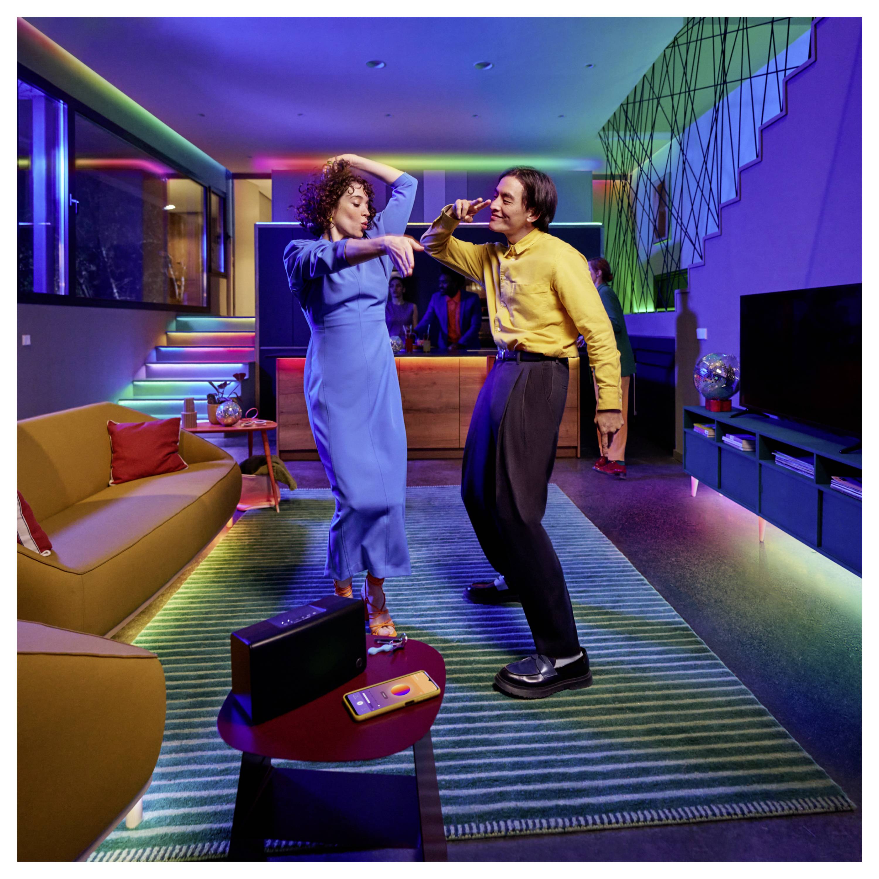 Une femme en robe bleue et un homme en chemise jaune dansent joyeusement dans un salon moderne et coloré, avec un éclairage vif et une décoration géométrique.