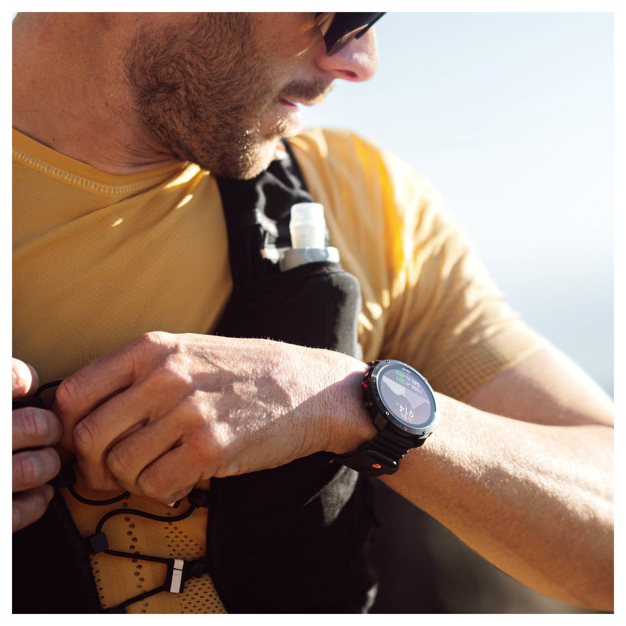 Un homme ajuste sa montre-bracelet noire tout en portant une chemise jaune et des lunettes de soleil. Il a un sac d'hydratation sur la poitrine. L'arrière-plan est flou.