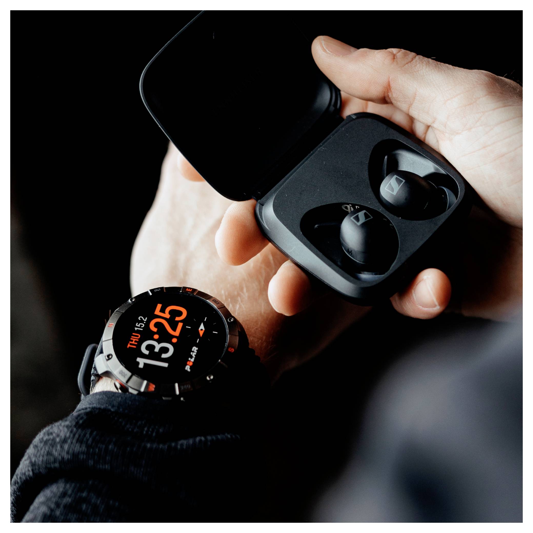 Une personne portant une montre connectée ouvre un boîtier noir d'écouteurs, révélant un écouteur. La montre affiche l'heure 13:25 et Jeu.