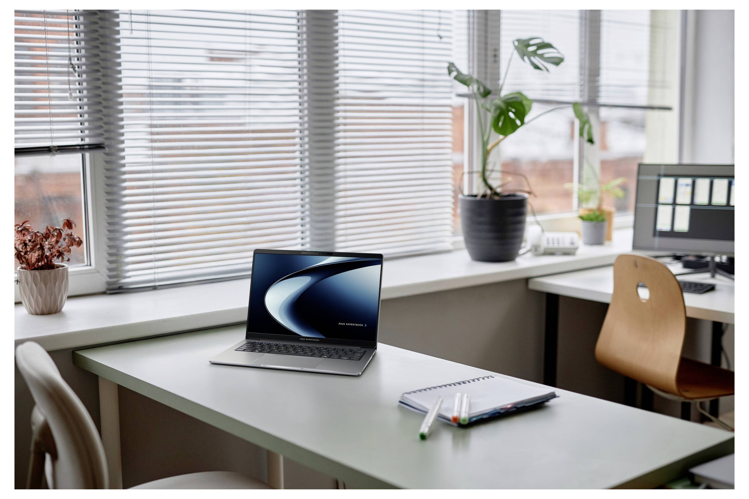 Un ordinateur portable moderne posé sur un bureau à côté de papeterie dans un espace de bureau lumineux avec de grandes fenêtres et des plantes, transmettant un environnement de travail productif.