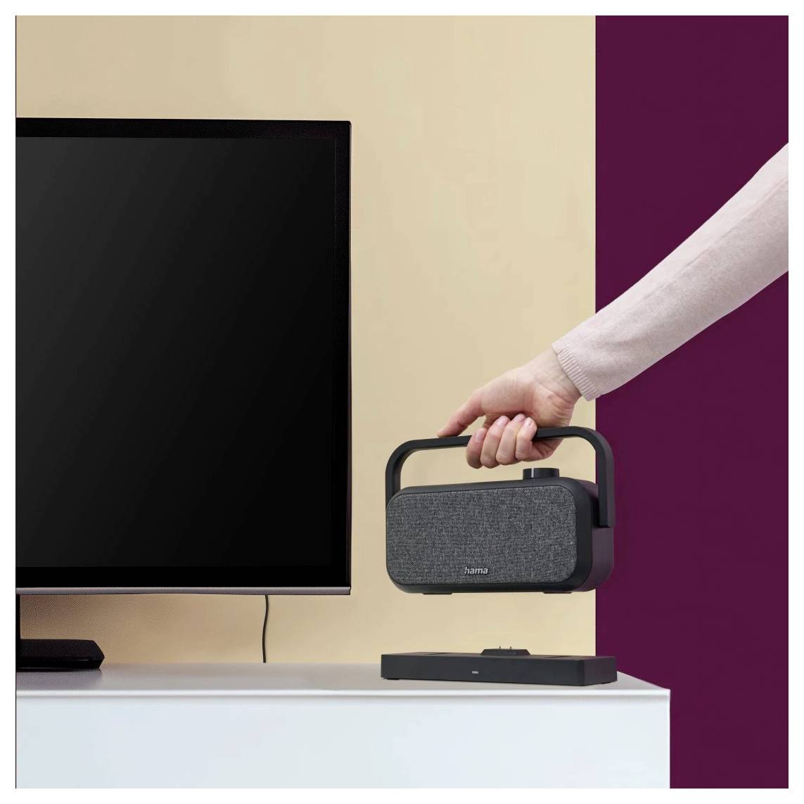 'Une personne soulève un haut-parleur sans fil d'une table. Un écran de télévision est partiellement visible à gauche. L'arrière-plan est marron et crème.'