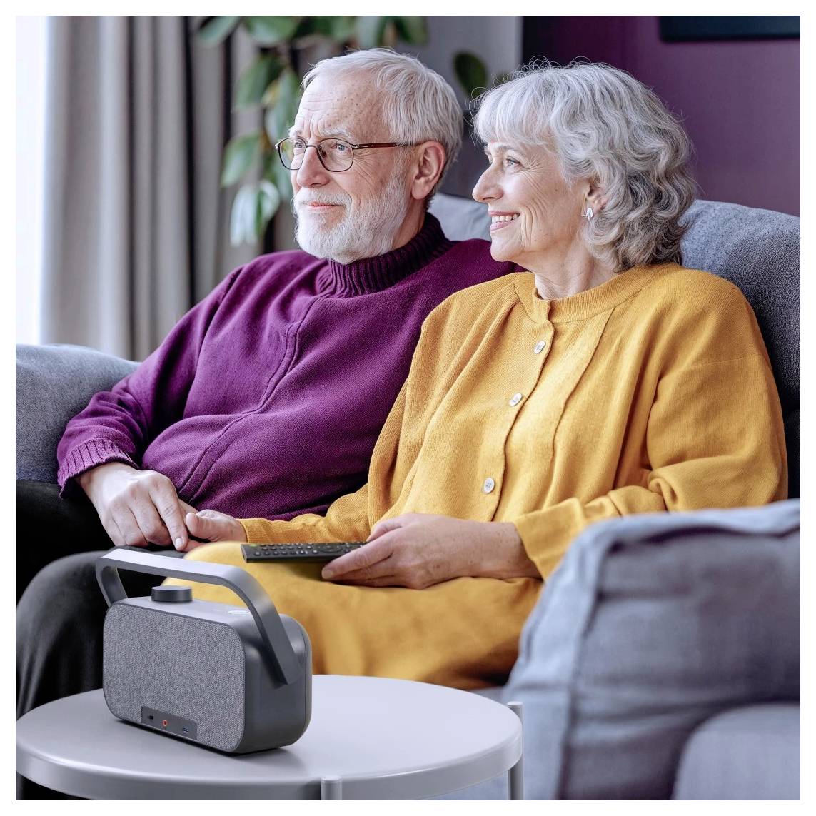 Un couple de personnes âgées est assis en souriant sur un canapé avec un haut-parleur sans fil posé sur une table au premier plan, suggérant un moment détendu et rempli de musique.
