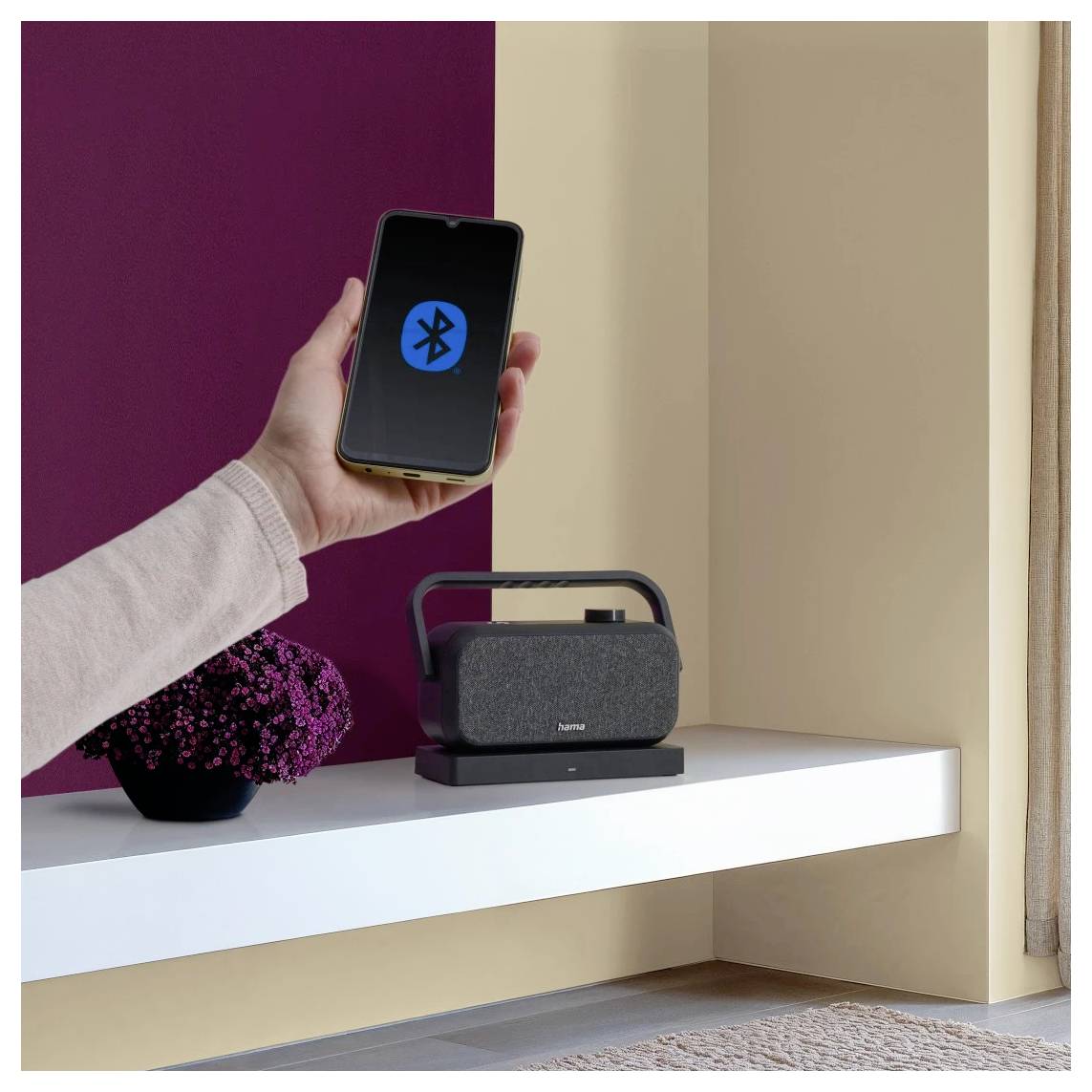Une main tenant un smartphone avec un symbole Bluetooth visible. À proximité, un haut-parleur portable est posé sur une étagère, à côté d'une petite plante.