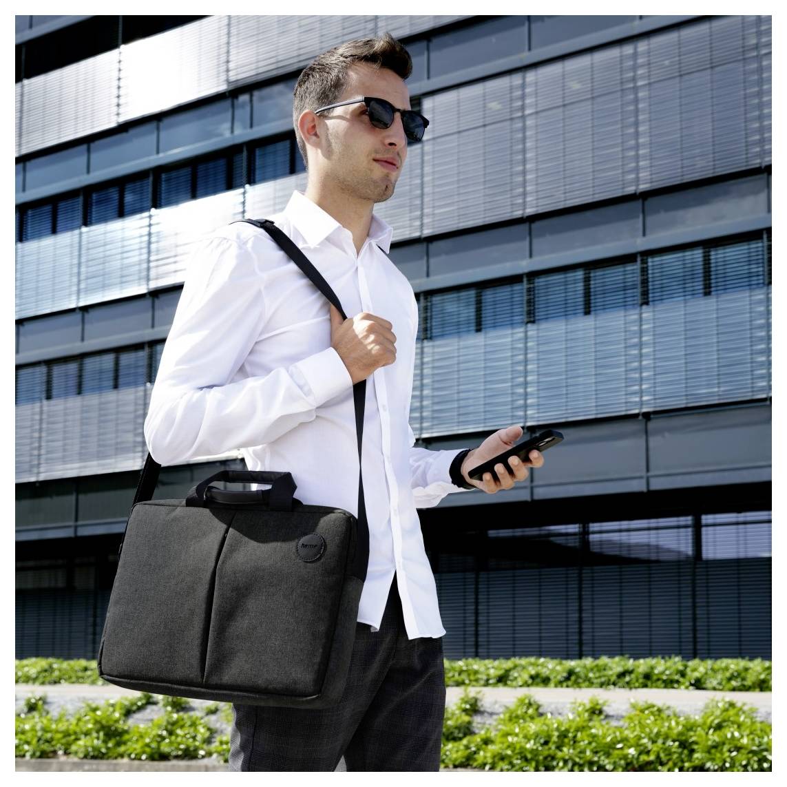 Un homme portant une chemise blanche et des lunettes de soleil tient un téléphone et une serviette noire, debout devant un immeuble moderne en verre.