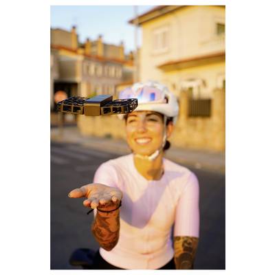 Une personne portant un casque de vélo et un t-shirt rose sourit tout en tendant la main vers un drone planant dans une rue ensoleillée.