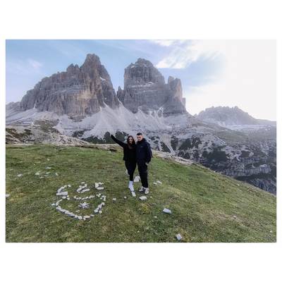Un couple se tient sur une colline herbeuse, avec une forme de cœur formée de pierres à leurs pieds. Des sommets de montagnes majestueuses s'élèvent en arrière-plan.
