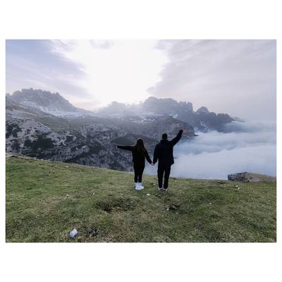 Un couple se tient sur un coteau herbeux, se tenant la main et faisant face à un panorama de montagnes et une vallée remplie de nuages sous un ciel lumineux.