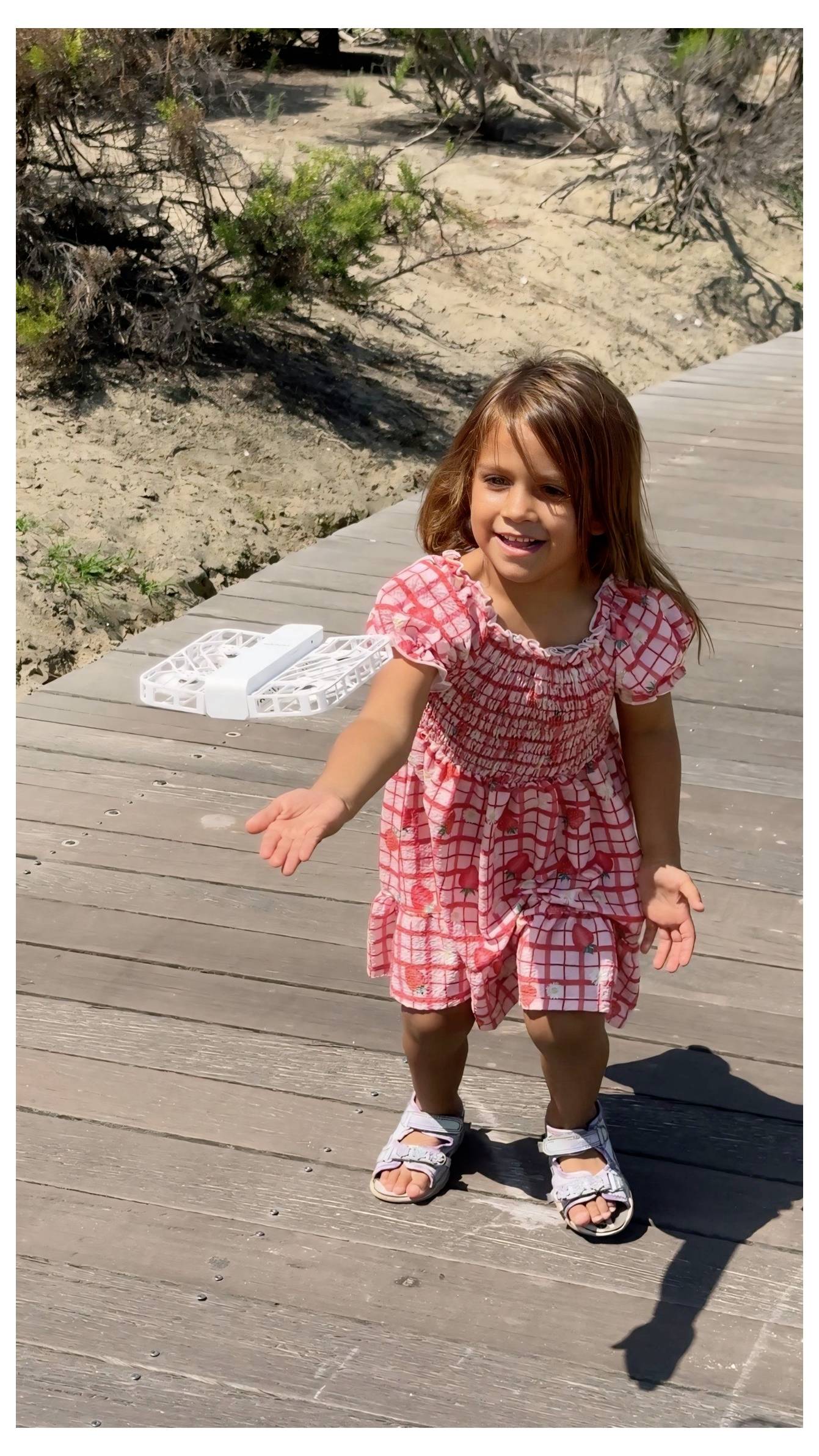 Une petite fille portant une robe rouge et blanche se tient sur une passerelle en bois, tendant les mains vers l'avant, avec de la verdure et du sable en arrière-plan.