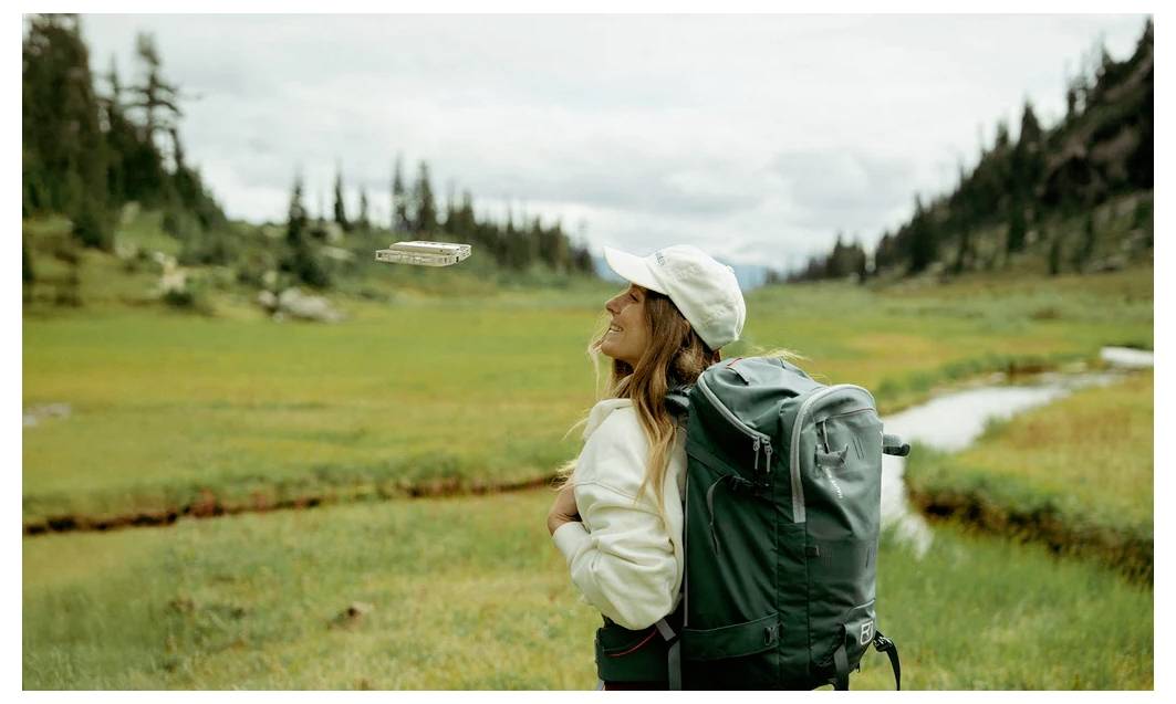 Une personne portant un sac à dos regarde au loin avec bonheur dans une prairie verdoyante avec des arbres et des montagnes.