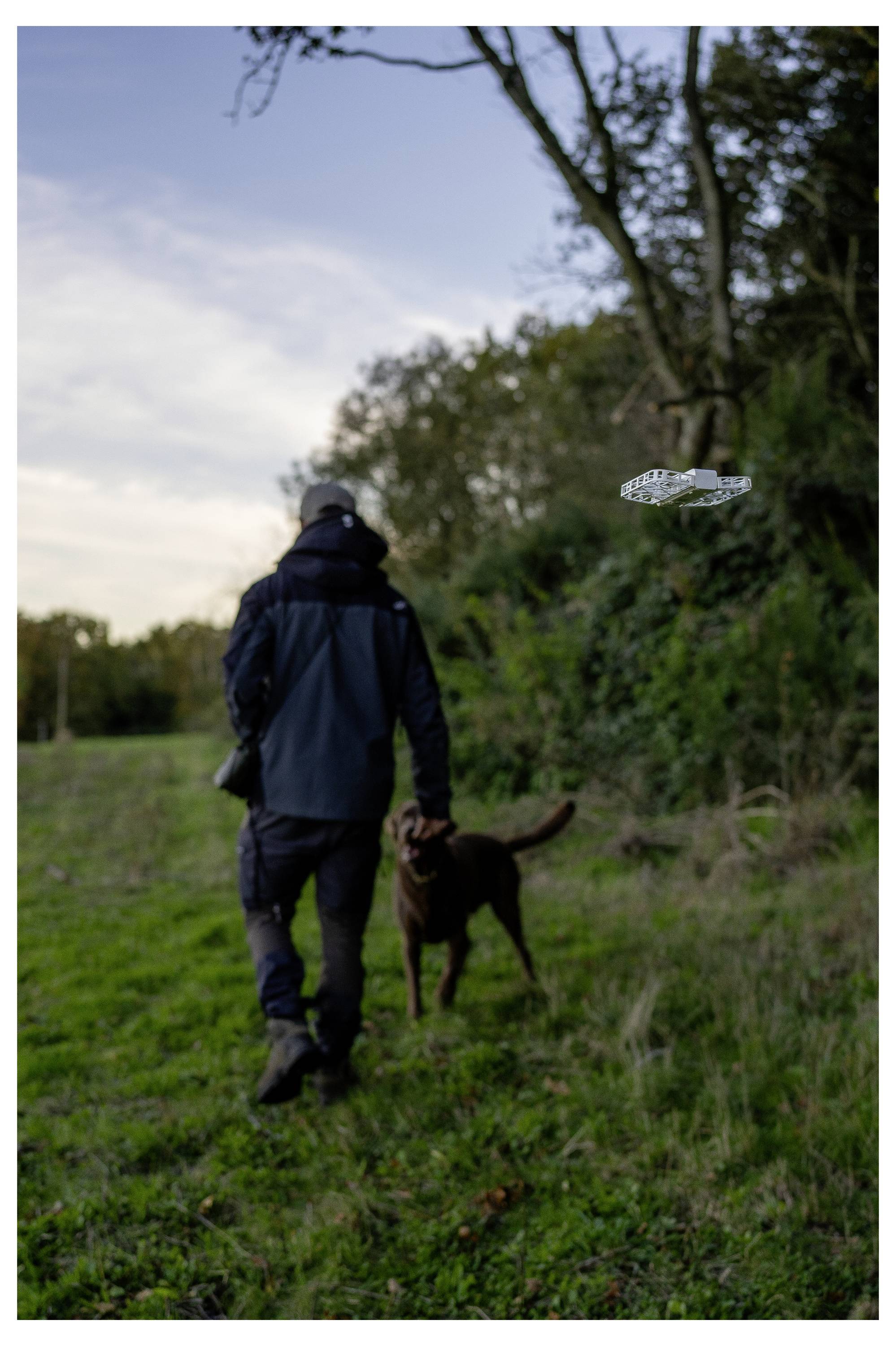 Une personne marche sur un sentier herbeux avec un chien brun, sous un ciel nuageux, tandis qu'un petit objet métallique brillant flotte à proximité.
