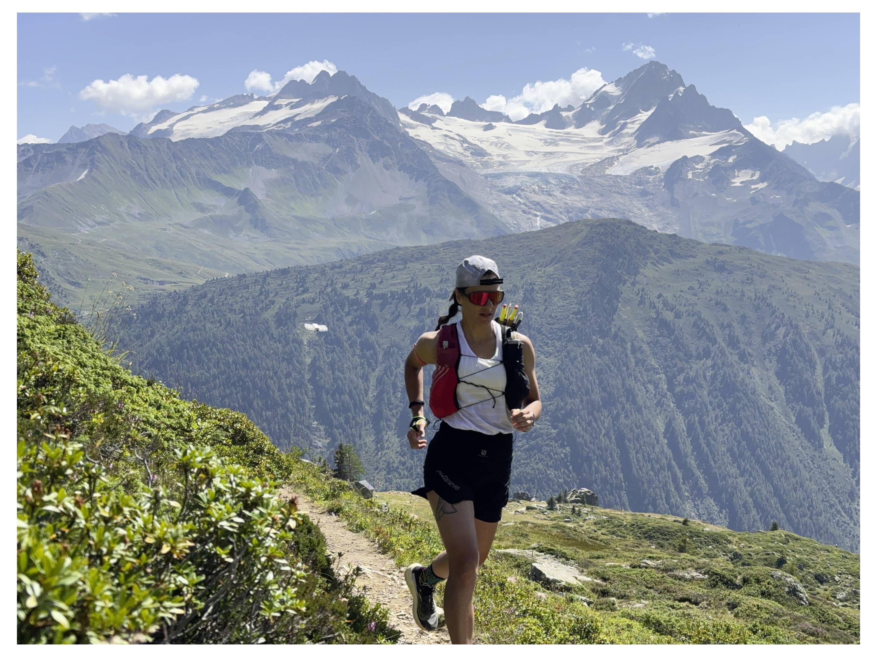 Une personne court sur un sentier de montagne entouré de verdure, avec des sommets enneigés en arrière-plan sous un ciel bleu clair.