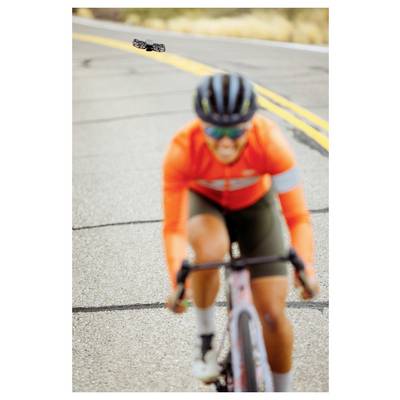Cycliste portant une veste orange et un short vert, roulant sur une route, avec un effet de flou, le revêtement et les lignes jaunes étant visibles.
