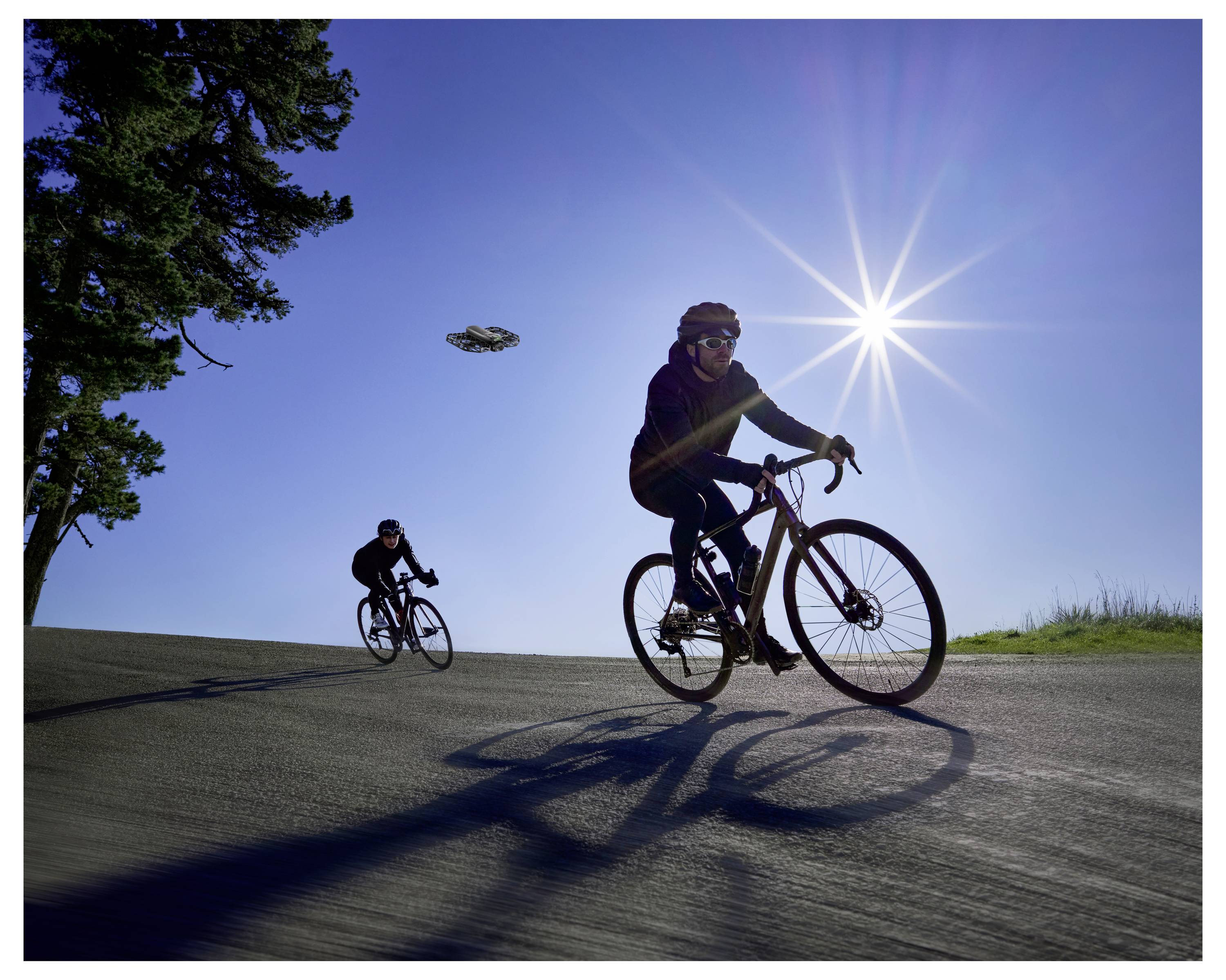 Deux personnes faisant du vélo en montée un jour ensoleillé, avec une silhouette d'arbres sur la gauche. Le soleil est éclatant dans le ciel dégagé.