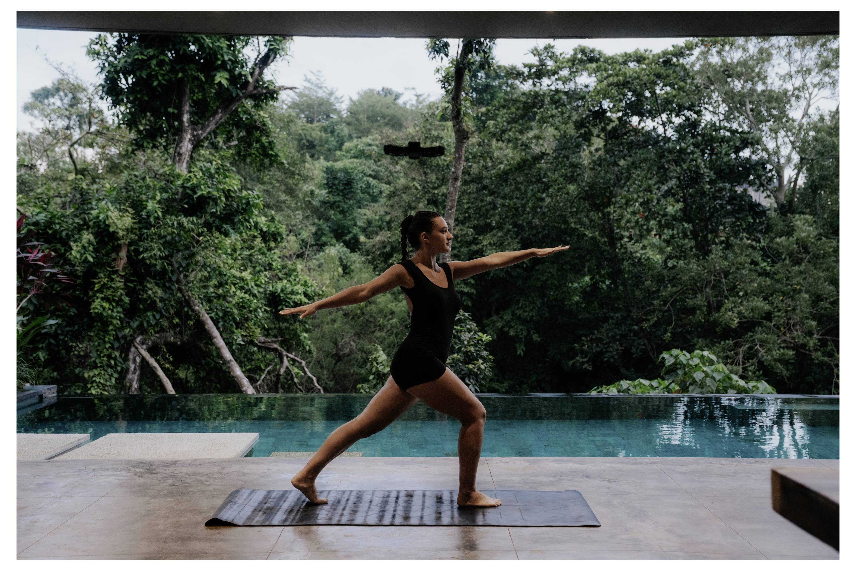Une personne pratique le yoga dans une posture de guerrier sur un tapis près d'une piscine à débordement, entourée d'arbres verts luxuriants dans un cadre extérieur serein.