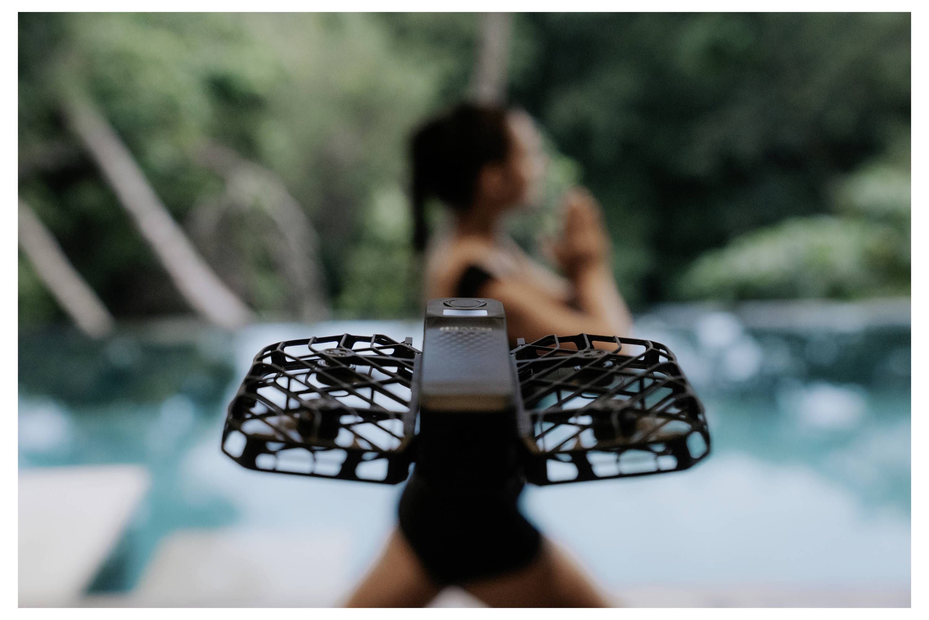 Une personne dans une posture de yoga près d'une piscine, avec un drone en vol au premier plan, et un arrière-plan verdoyant et luxuriant.