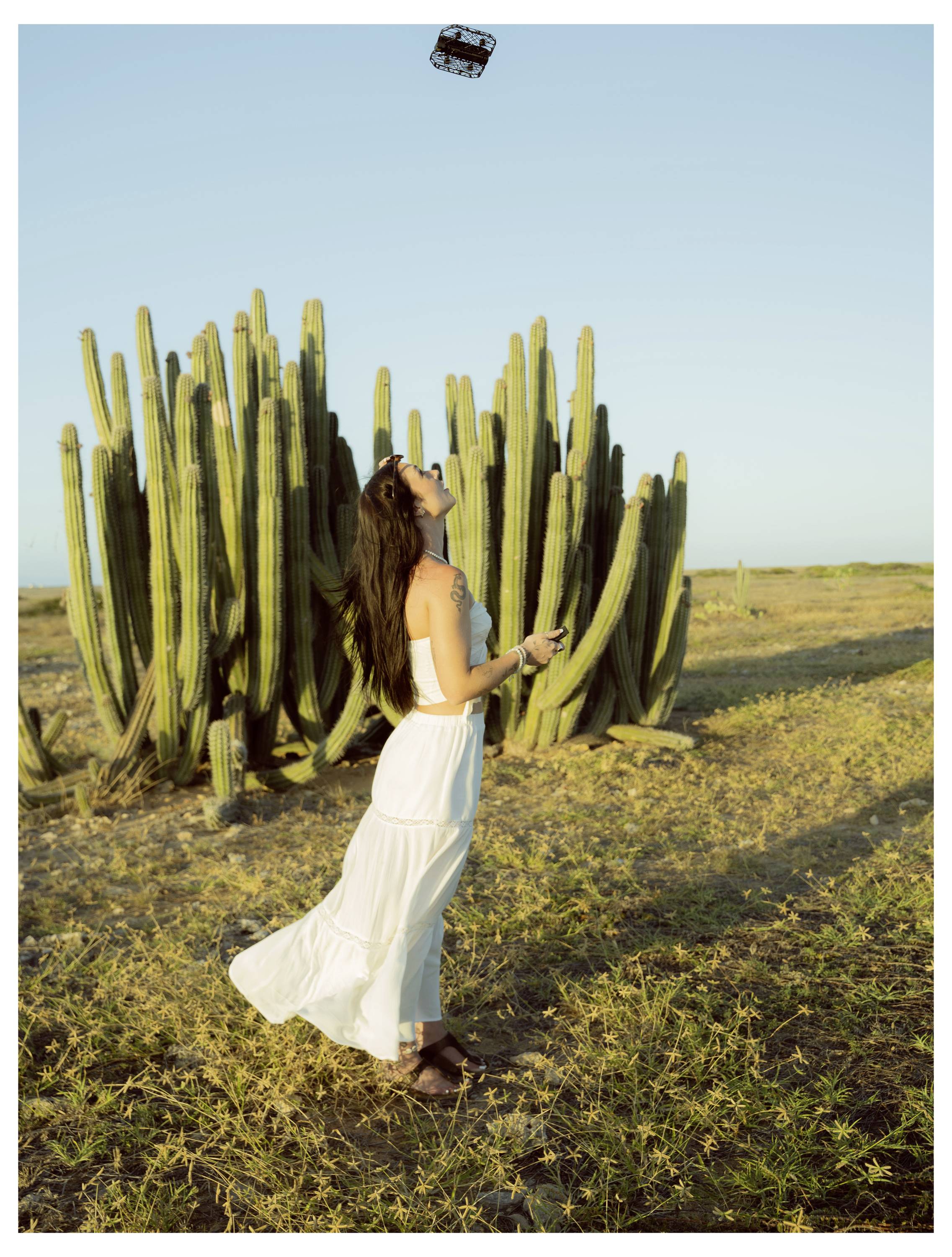 Une femme en robe blanche se tient dans un paysage désertique, jetant un petit objet en l'air. De grands cactus se dressent en arrière-plan sous un ciel dégagé.