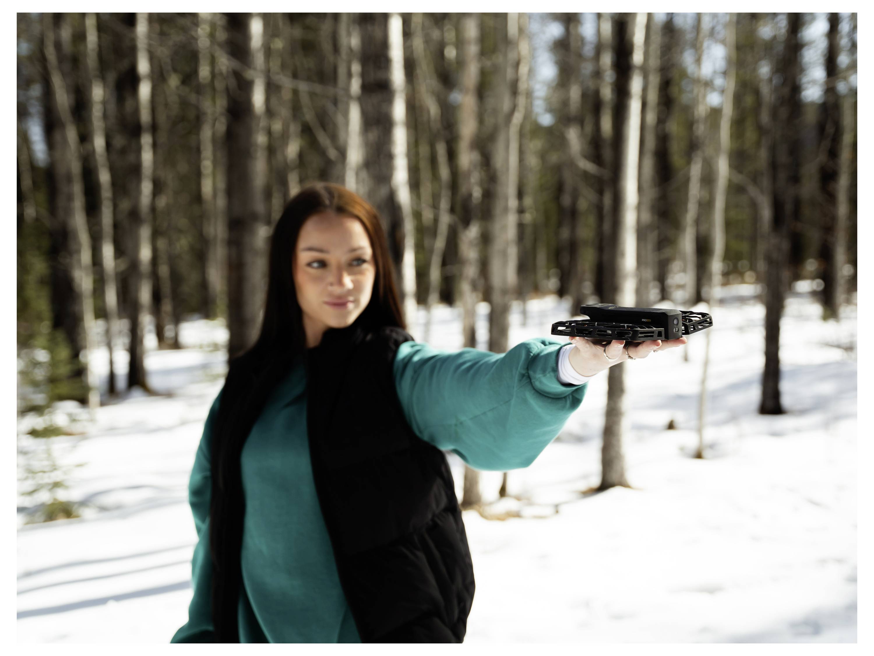 Une personne dans une forêt enneigée tient un drone, prête à le lancer. Le soleil brille, projetant sa lumière sur la neige et les arbres.
