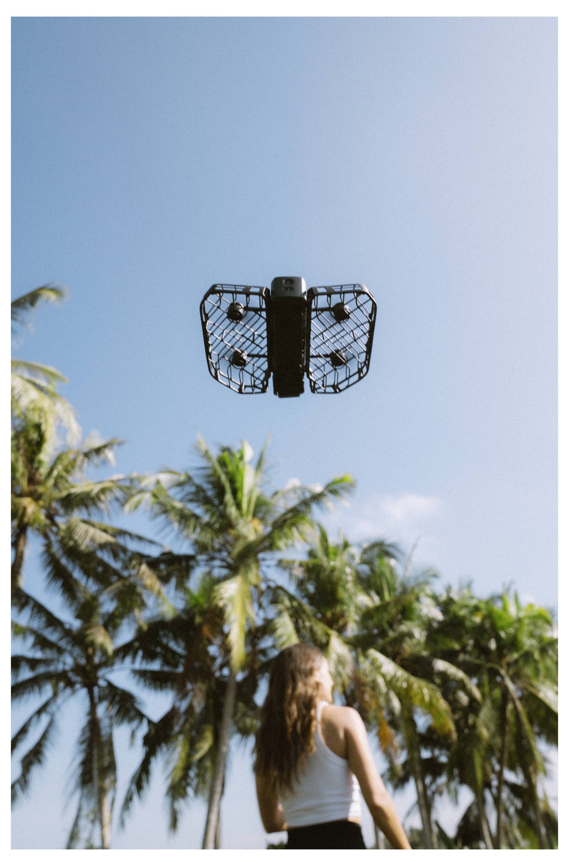 Une femme se tient sous des palmiers, regardant vers le haut un drone volant au-dessus d'elle contre un ciel bleu clair.