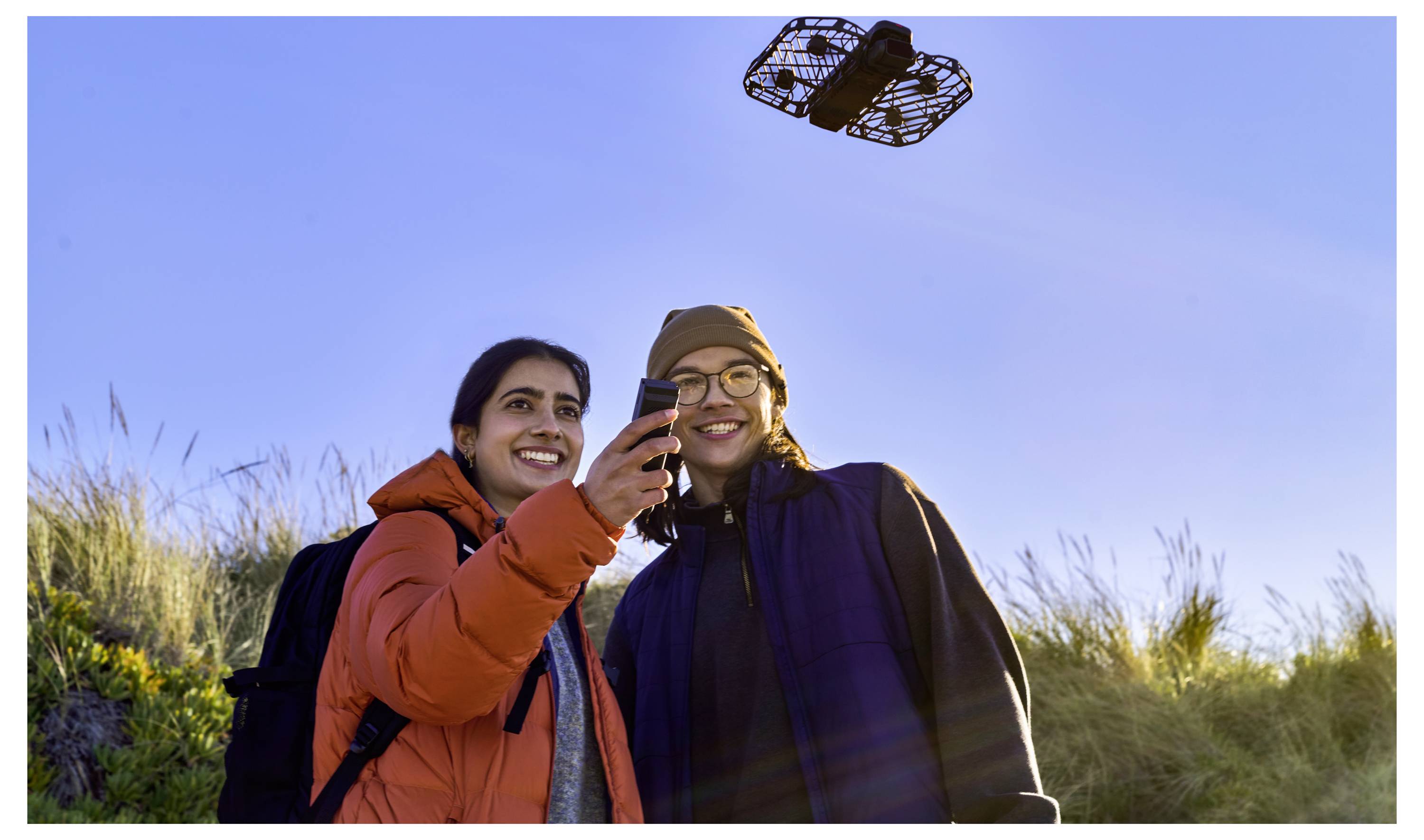 Deux personnes vêtues de tenues d'extérieur sourient tout en contrôlant un petit drone volant au-dessus de dunes herbeuses sous un ciel bleu dégagé.