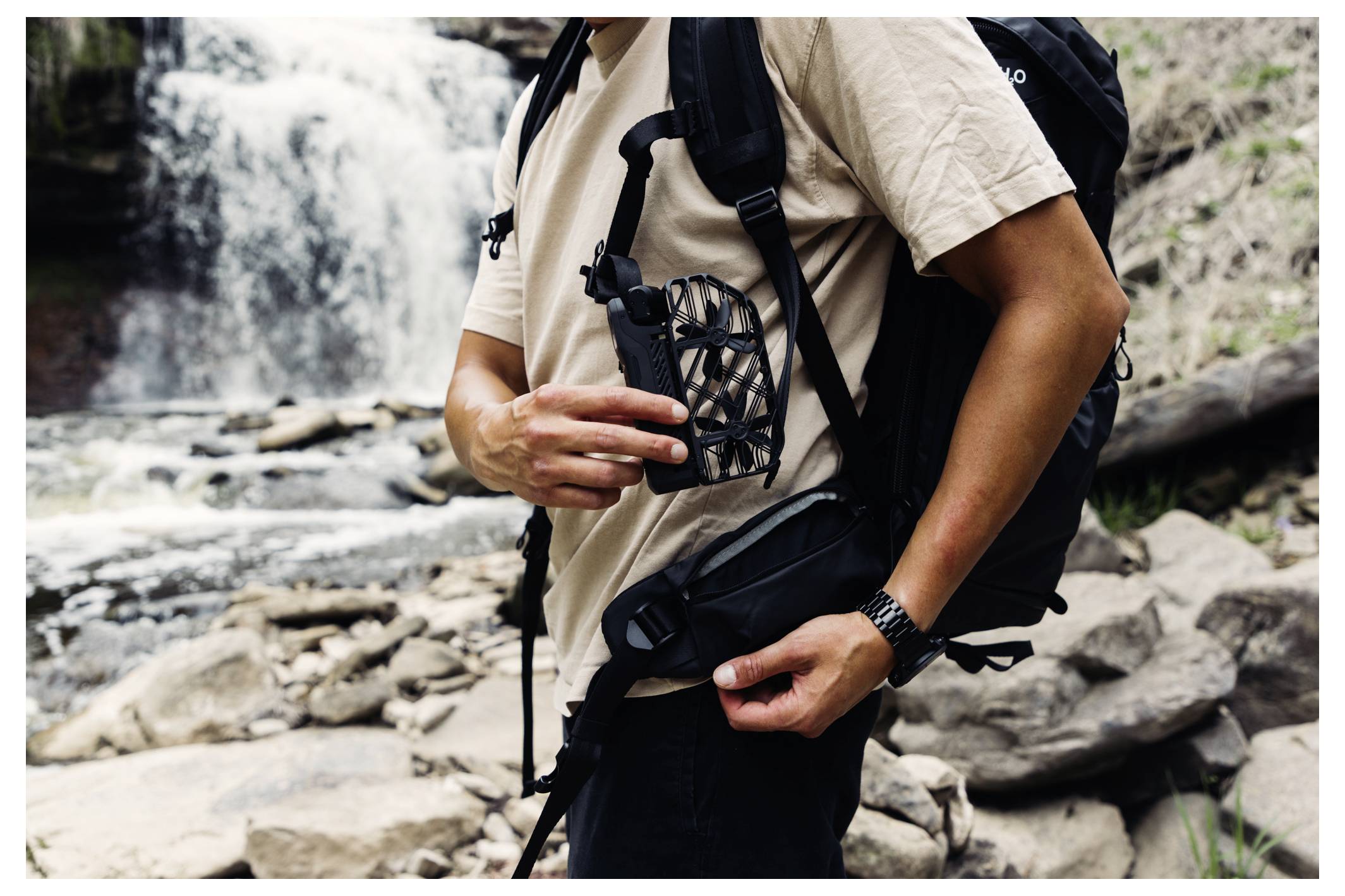 Un randonneur près d'une cascade, tenant un ventilateur portable. Il porte un sac à dos et une chemise beige, debout sur un terrain rocailleux.