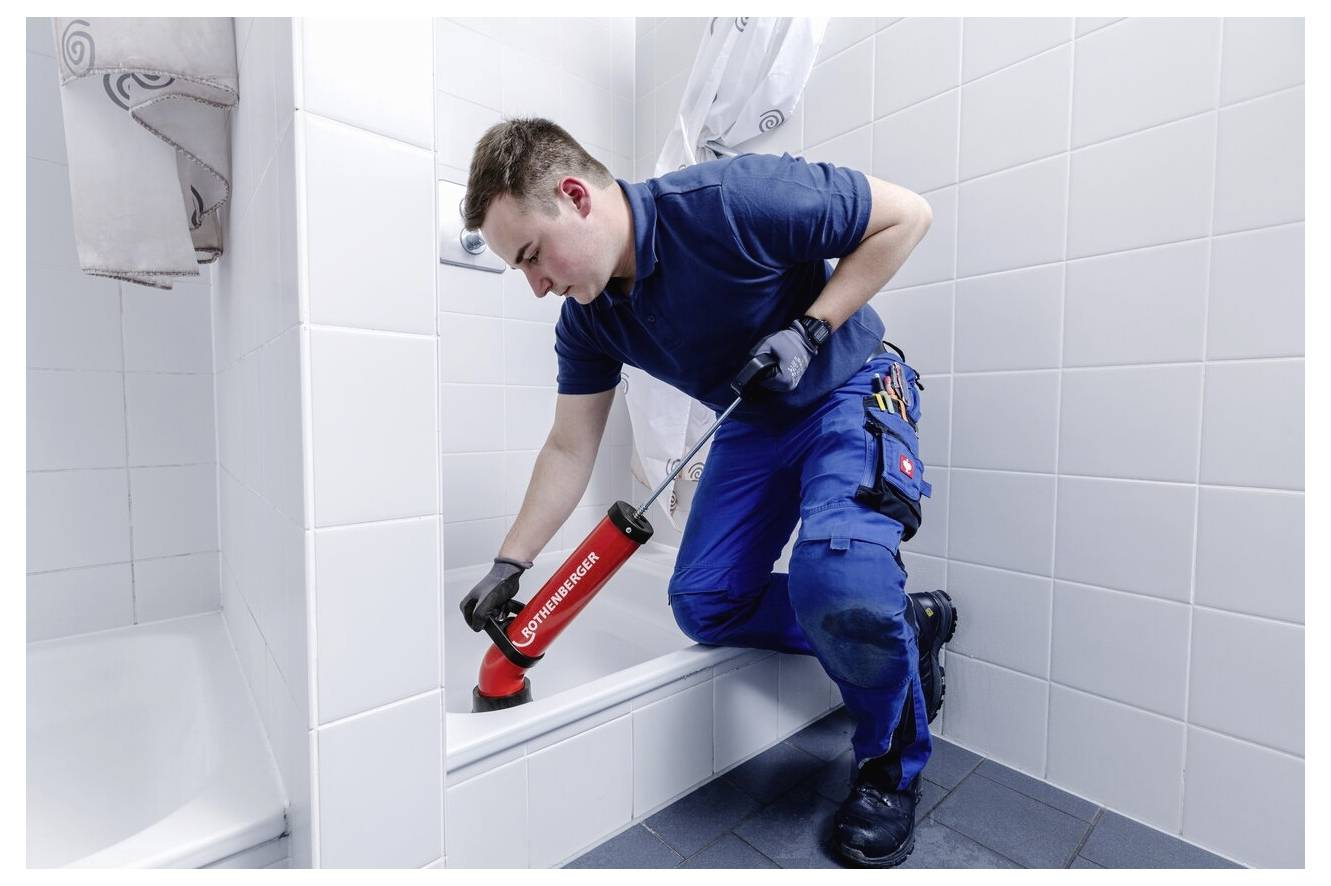 Un plombier portant une salopette bleue utilise un pistolet à silicone pour sceller le bord d'une baignoire dans une salle de bain carrelée, en se concentrant sur la précision.