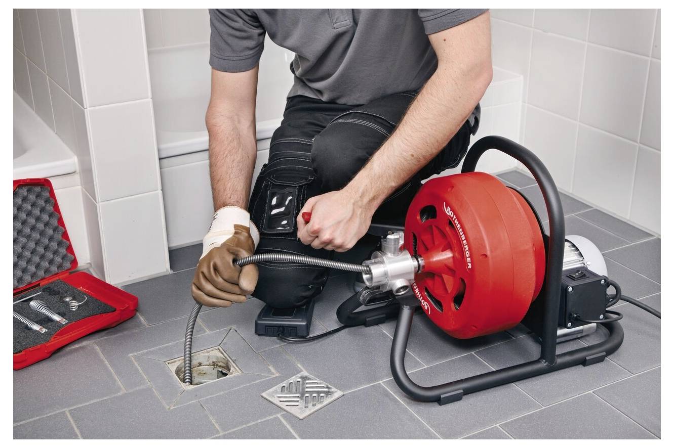 Un plombier utilise un déboucheur mécanique pour déboucher un siphon de sol de salle de bain. Une boîte à outils est ouverte à proximité, montrant divers instruments.