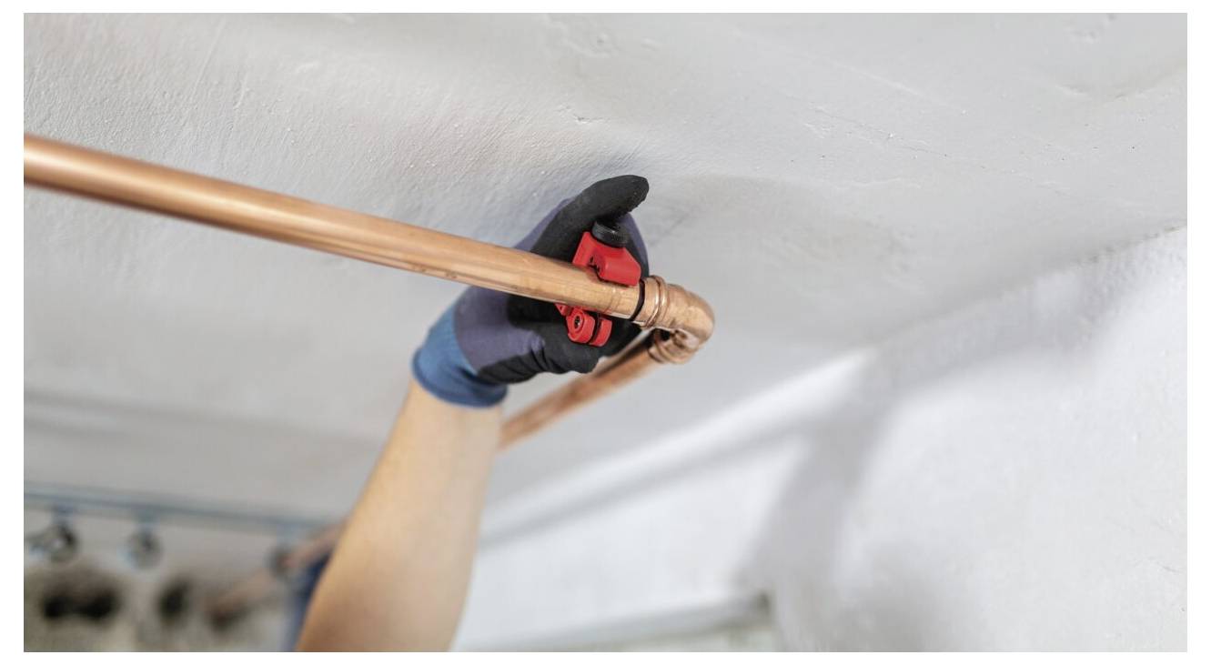 Une personne portant des gants utilise un coupe-tube pour couper un tuyau en cuivre fixé à un plafond, ce qui indique des travaux de plomberie ou de rénovation.