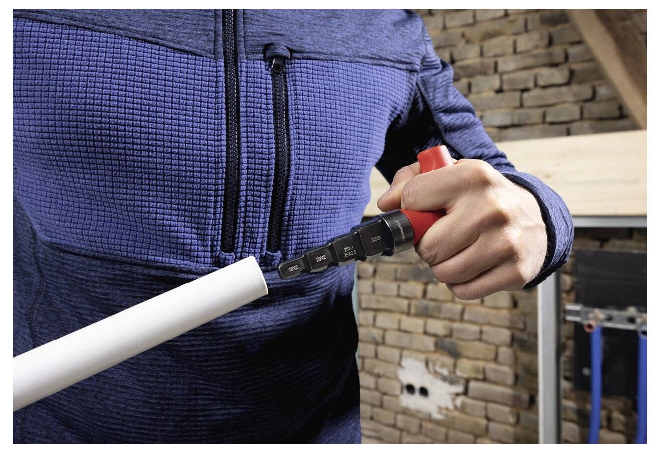 Une personne portant une veste bleue utilise un outil pour couper un tuyau blanc à l'intérieur, près d'un mur de briques en cours de rénovation.