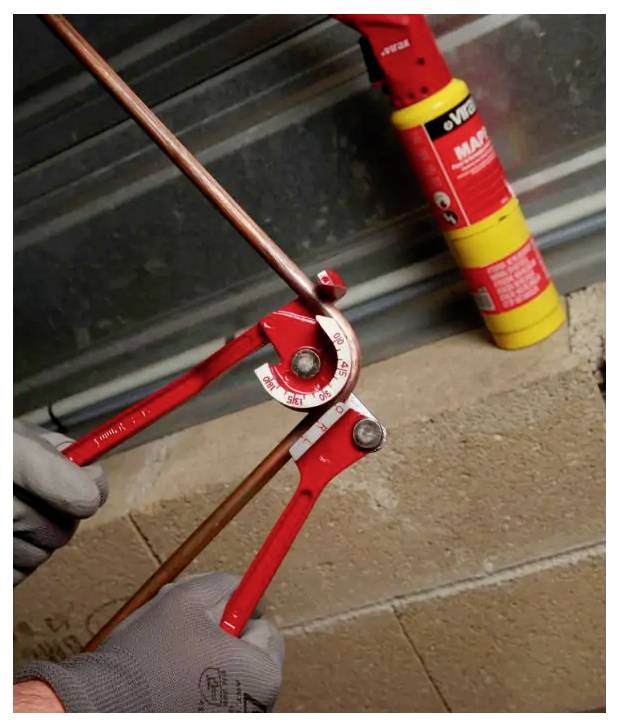 Des mains gantées utilisent un cintre de tuyauterie rouge pour plier un tube en cuivre, avec un extincteur en arrière-plan.