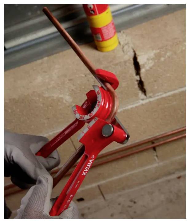 Main gantée utilisant un cintreuse de tuyau rouge pour plier un tuyau en cuivre contre un mur de briques.