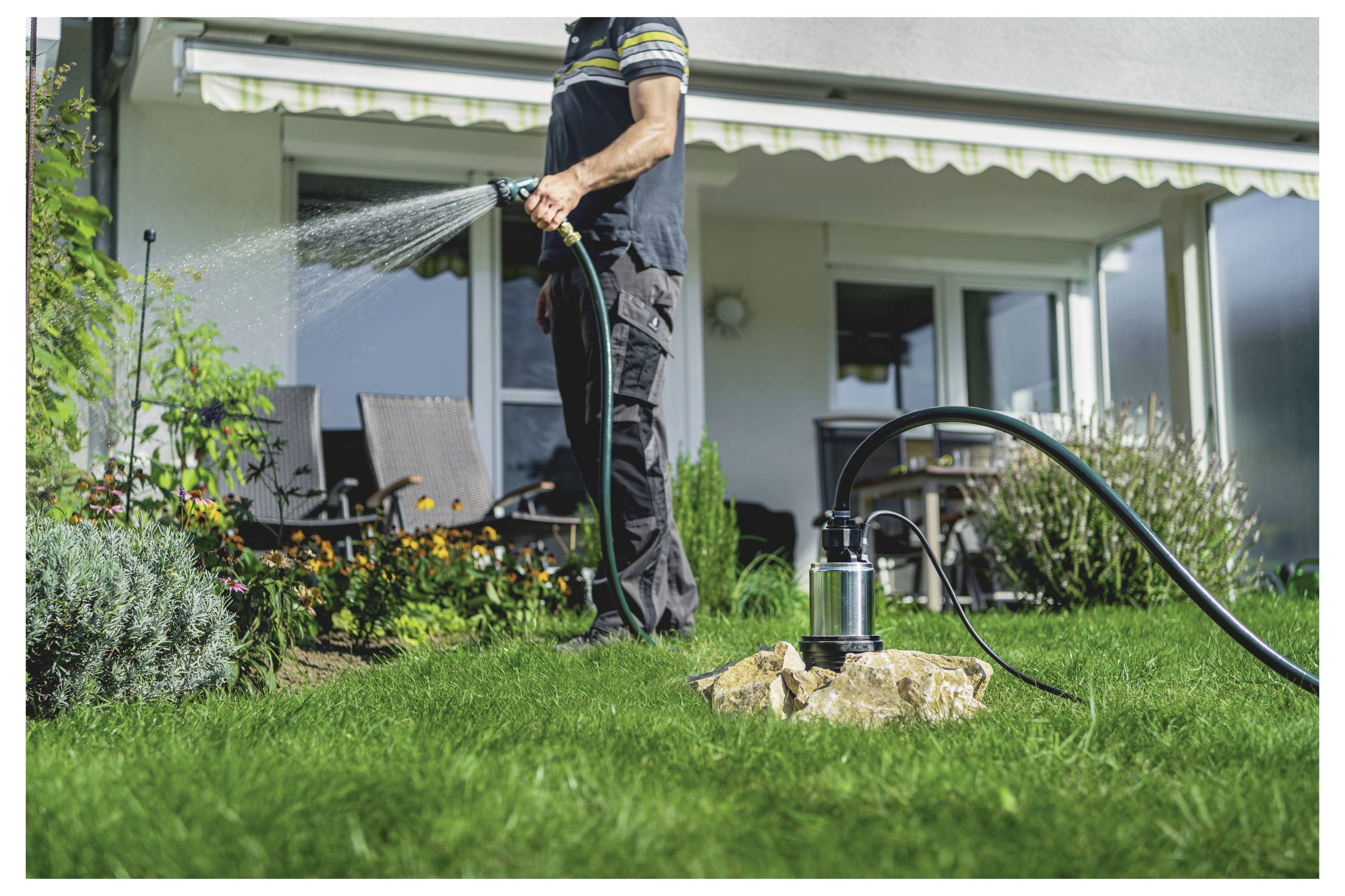 Une personne arrose un jardin avec un tuyau d'arrosage, debout sur une pelouse verte. Du mobilier de terrasse et des plantes en fleurs sont en arrière-plan.