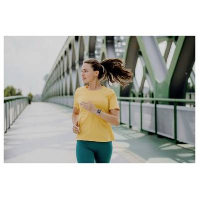 Une femme portant un t-shirt jaune fait du jogging sur un pont vert par une journée claire, regardant droit devant elle avec une détermination concentrée.