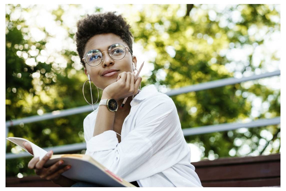 Une personne assise en extérieur, portant des lunettes et tenant un carnet et un stylo, a l'air pensif avec des arbres en arrière-plan flou.