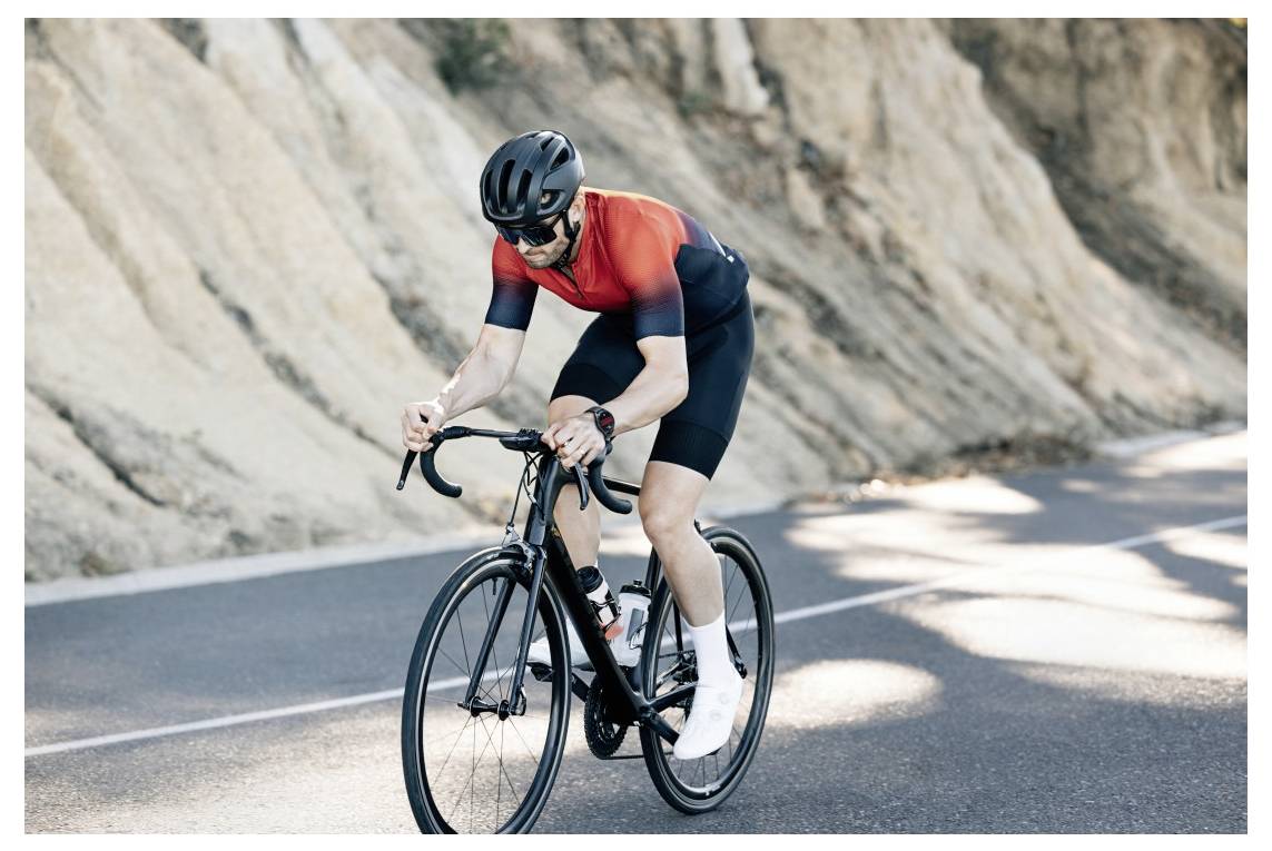Un cycliste portant une tenue rouge et noire roule sur un vélo de route sur une route de montagne sinueuse, éclairée par le soleil et bordée de rochers.