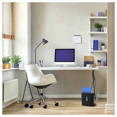 Un bureau à domicile moderne avec un bureau, un ordinateur, une lampe, une chaise, des étagères avec des plantes et des livres, et un destructeur de documents sur le sol. La lumière inonde la pièce.