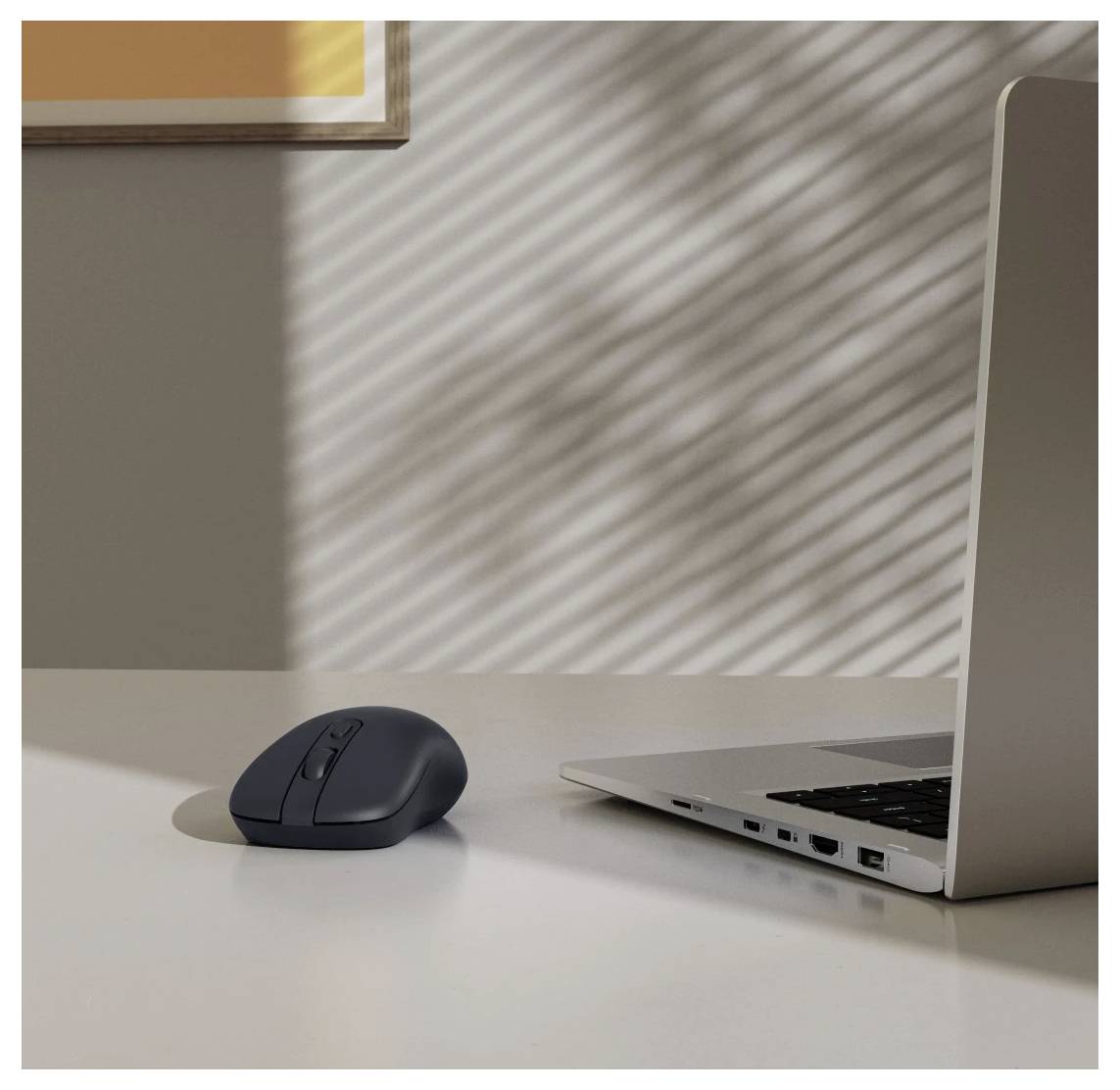 « Une souris d'ordinateur à côté d'un ordinateur portable argenté ouvert sur une table blanche ; la lumière du soleil crée des ombres rayées sur le mur en arrière-plan. »