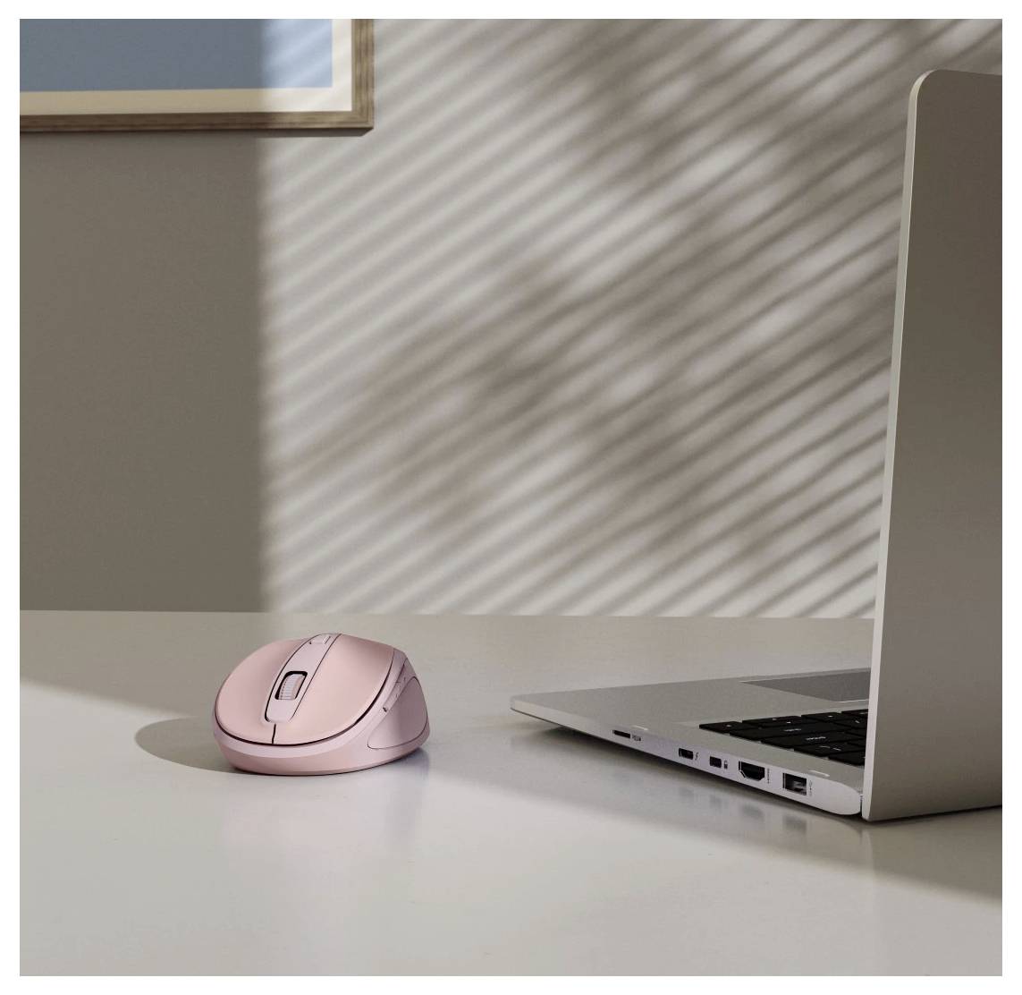 Une souris d'ordinateur rose et un ordinateur portable ouvert sur un bureau, avec la lumière du soleil projetant des ombres striées à travers les stores de la fenêtre.