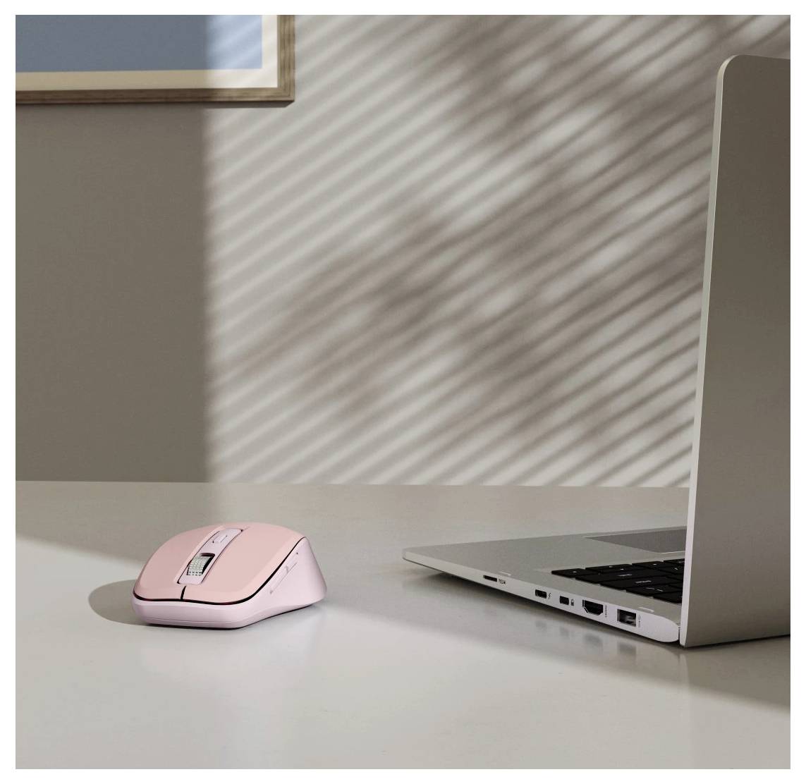 Une souris d'ordinateur rose et un ordinateur portable ouvert sont posés sur un bureau blanc. La lumière du soleil projette des ombres striées des stores sur le mur et le bureau.