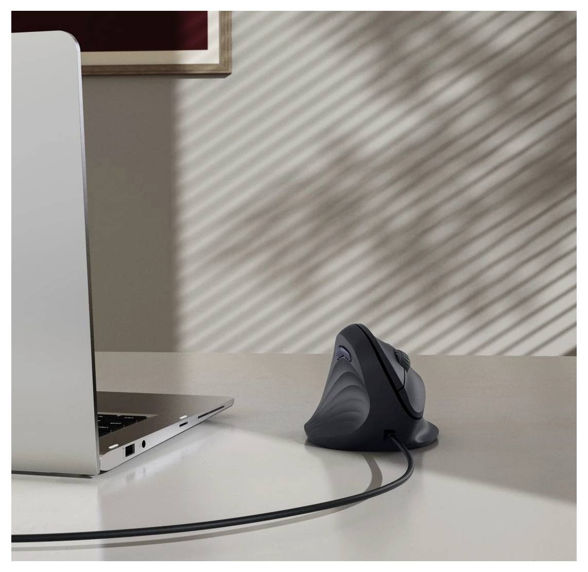 Un ordinateur portable sur un bureau à côté d'une souris ergonomique verticale. La lumière du soleil crée des ombres striées sur le mur, rehaussant l'ambiance de l'espace de travail.