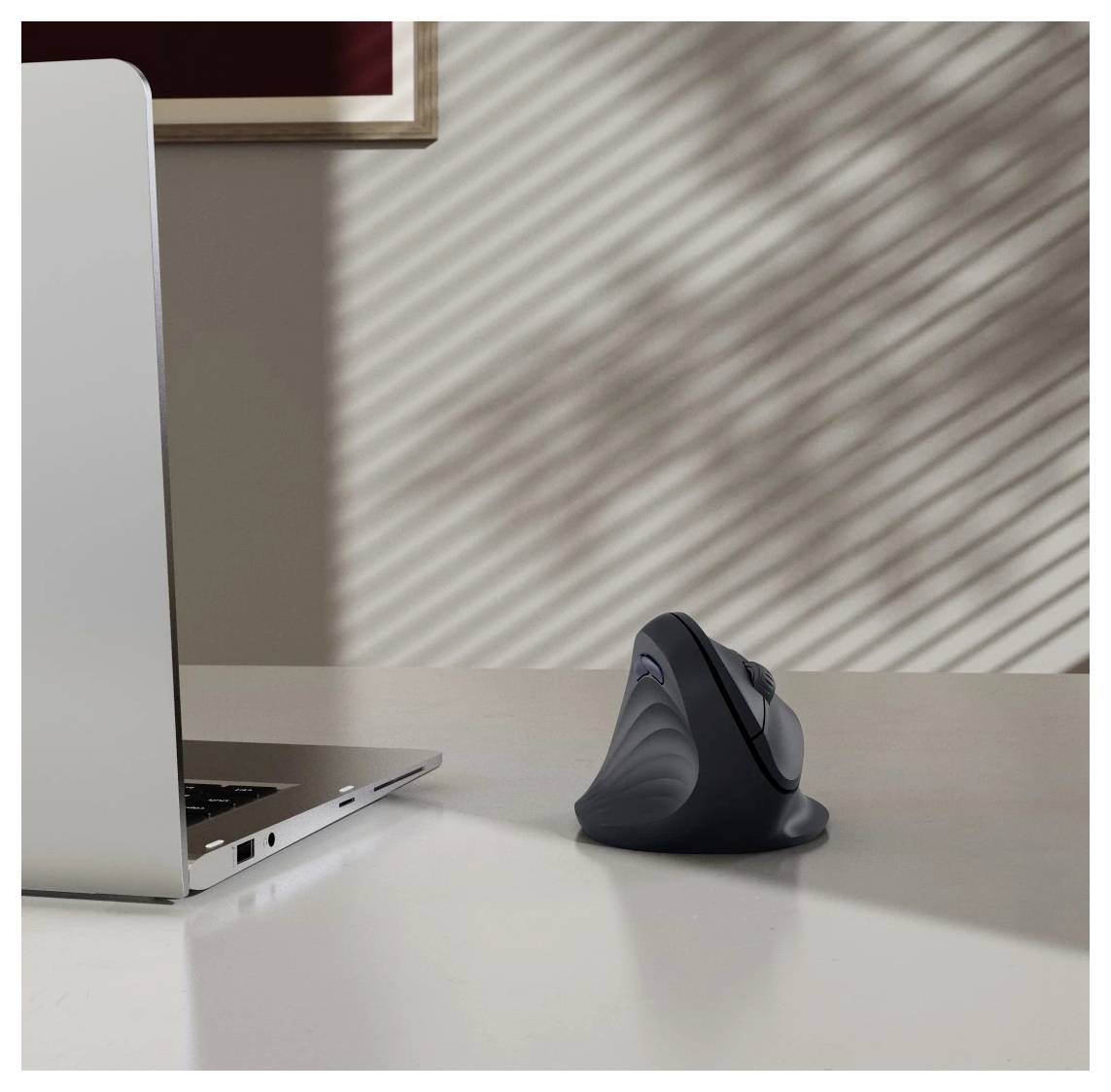 Un ordinateur portable et une souris ergonomique sur une table blanche sont illuminés par une lumière douce et striée passant à travers les stores.