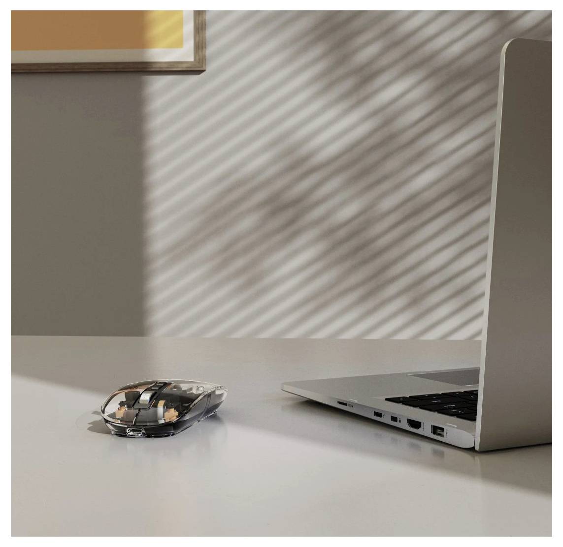 Une souris d'ordinateur élégante et transparente à côté d'un ordinateur portable argenté sur un bureau baigné de soleil, avec des motifs d'ombres projetés par des stores sur le mur.