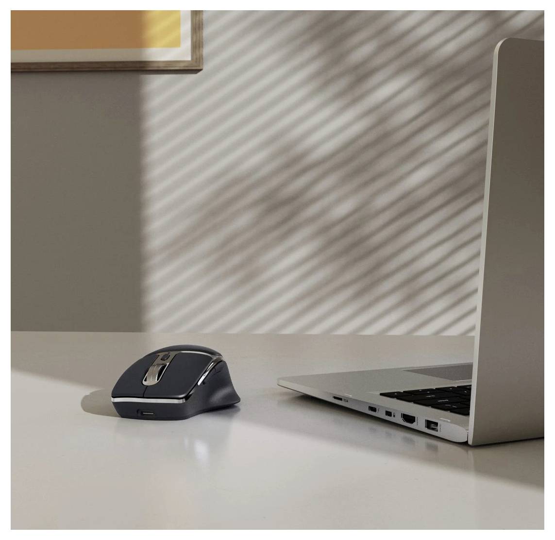 Une souris sans fil est posée sur un bureau blanc à côté d'un ordinateur portable ouvert, avec la lumière du soleil projetant des ombres striées sur la surface.