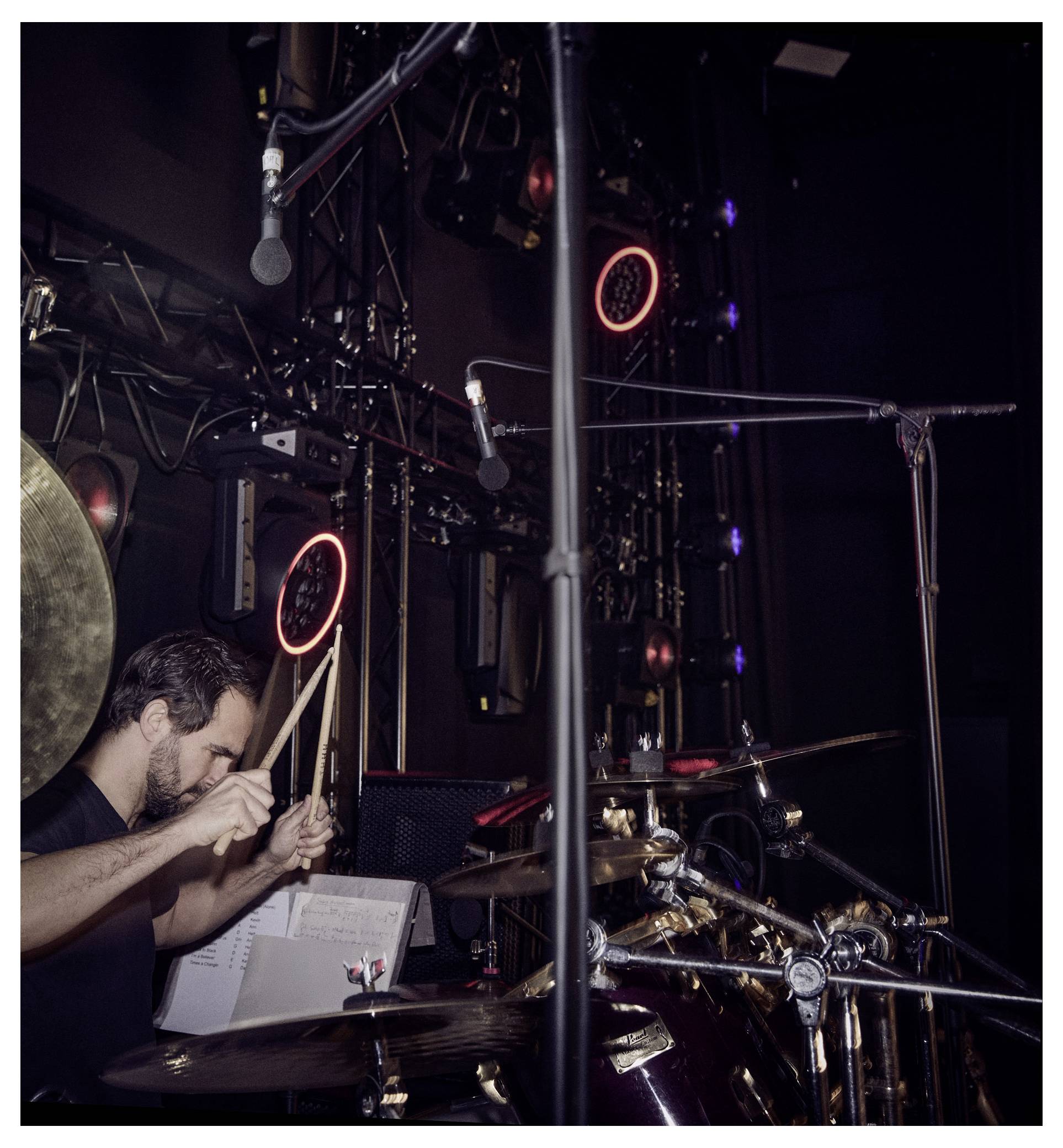 Batteur sur scène jouant une batterie, entouré de microphones et de lumières de scène.
